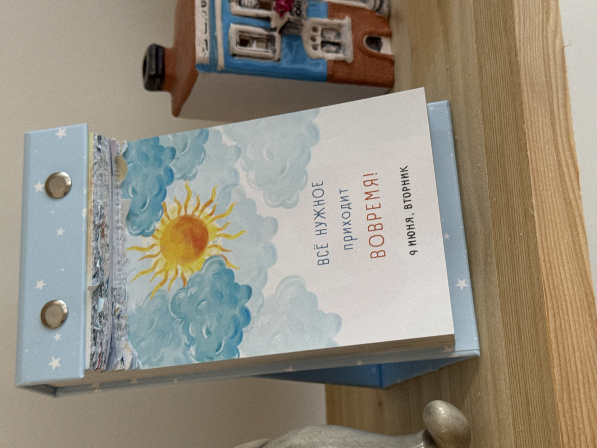 The image shows a close-up of a desk calendar with a watercolor illustration of a sun and clouds. Text on the calendar reads "Всё нужное приходит вовремя!" (Everything needed comes in time!) and "9 июня, вторник" (June 9, Tuesday). The calendar is light blue with white stars and has a wire binding. To the right of the calendar is a small, decorative house figurine, painted blue and brown with white trim. The objects are placed on a light-colored wooden shelf. There are no people or animals visible in the image. The location is not specified within the image content.