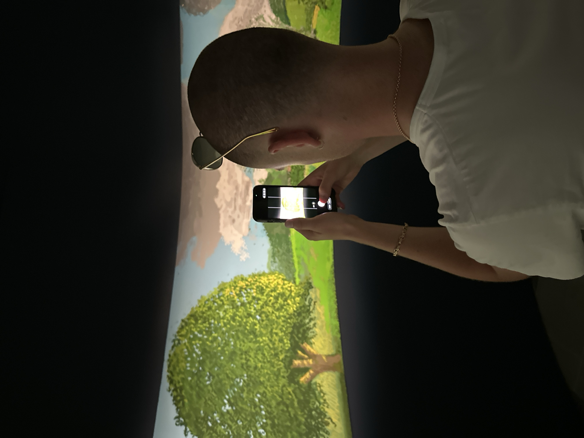 A person with a shaved head is holding a smartphone and taking a picture of a large, curved, animated mural depicting a bright green tree in a landscape. The person is wearing a white t-shirt, a gold chain necklace, and sunglasses on their head. The scene appears to be indoors, possibly in a museum or gallery, with dark surroundings contrasting with the illuminated mural. The location is not specified in the image, and therefore, I cannot provide information about the city of London.