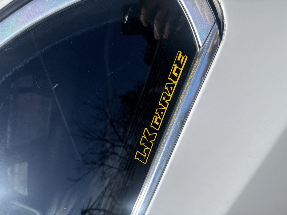 This image depicts a close-up of the rear driver's side window and adjacent body panel of a light grey or silver vehicle, observed in Daskal-Atanasovo, Bulgaria. Affixed to the lower portion of the dark-tinted window is a yellow adhesive decal spelling "LK GARAGE" in capitalized block letters with a black outline. The window surface exhibits multiple minor scratches, dust, and streaking. Reflected in the glass are bare tree branches against a bright, clear sky, indicating an outdoor, daytime setting. A partial reflection of a hand holding a dark-colored smartphone, likely the device capturing the image, is visible in the lower right corner of the window. A chrome or silver trim piece separates the window from the vehicle's body panel, which appears clean and unblemished within the frame. No individuals are directly visible.