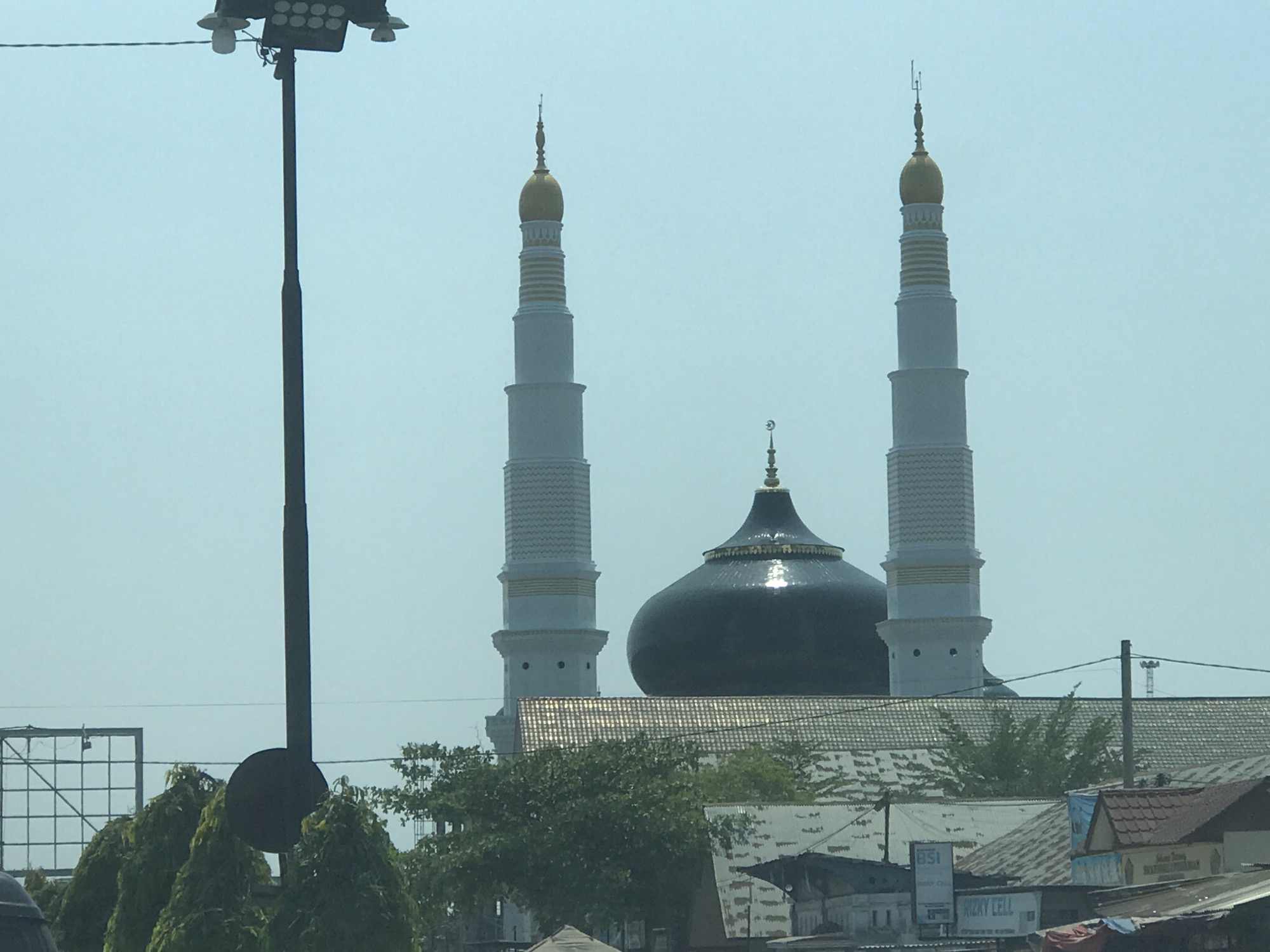 The image presents a view of the Baiturrahman Grand Mosque located in Keudematangglumpangdua, Indonesia. Two white minarets with gold finials flank a dark-colored central dome, which is surmounted by a crescent moon symbol. The mosque is partially obscured by trees and the roofs of buildings, giving the impression of being integrated into the urban landscape. A tall, dark-colored utility pole with multiple lights is visible in the foreground, obstructing some of the view. The overall environment suggests a daytime scene with a slightly hazy atmosphere.