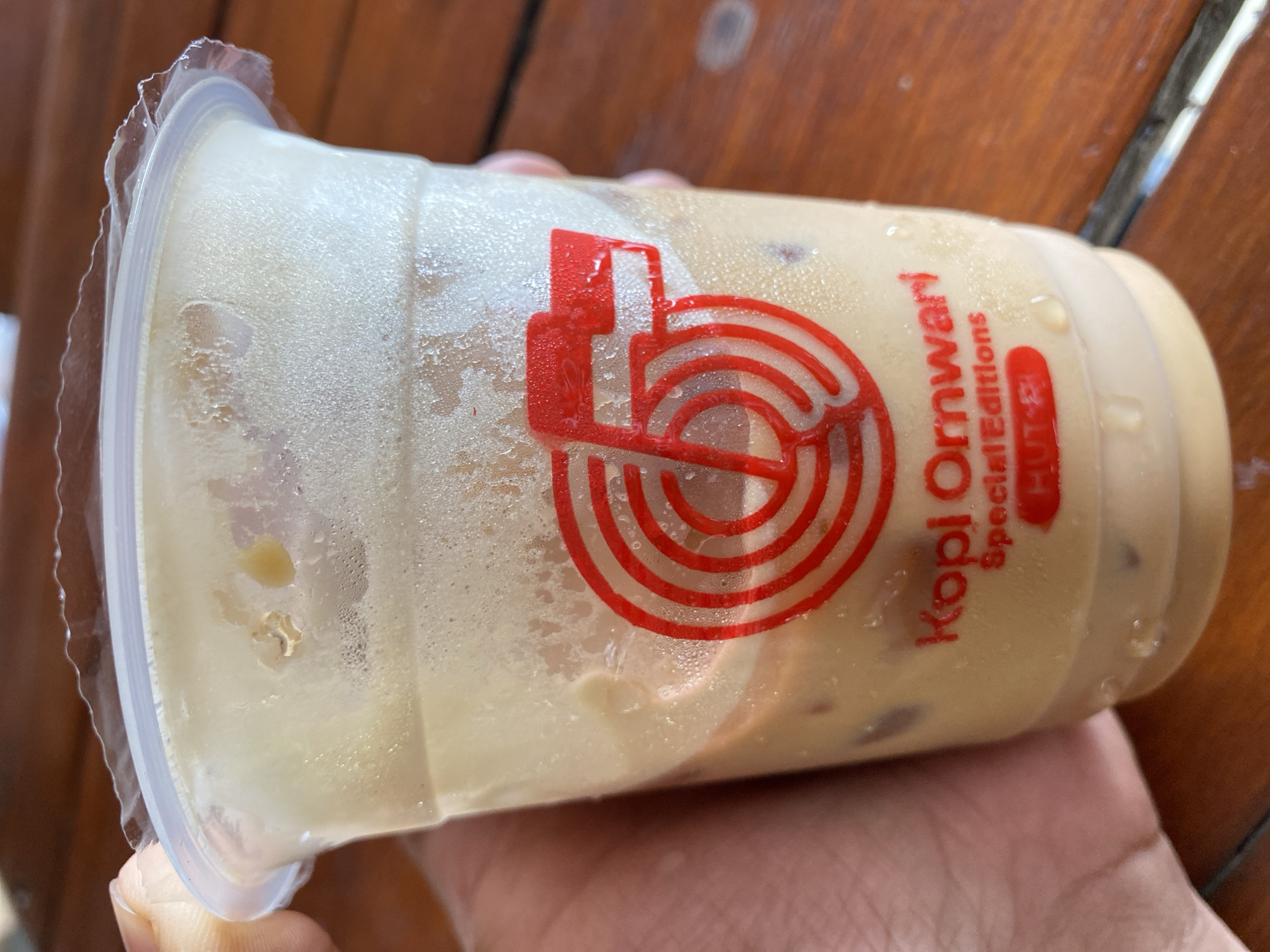 A close-up shot shows a hand holding a clear plastic cup filled with iced coffee with milk. The cup has a red logo with the number "5" and the text "Kopi Orwari Special Editions" printed on it. The cup is placed on a wooden table. The background is blurred, but appears to be outdoors, possibly in Bireuenmeunasahdayah, Indonesia, based on the mention of the city in the prompt. There is no visible action or interaction depicted.