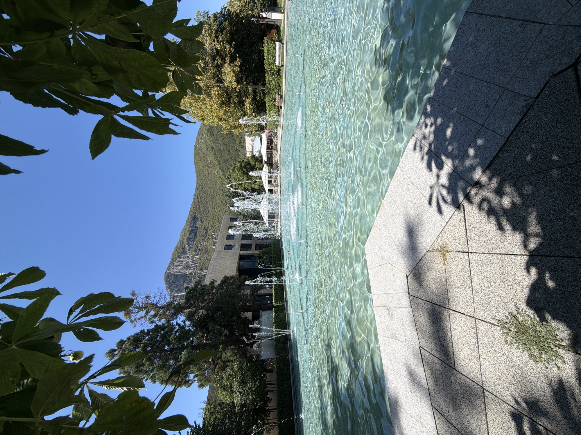 The image depicts a large, shallow fountain with multiple water jets creating a lively display. The water is clear and reflects the bright sunlight, creating shimmering patterns on the surface. The fountain is surrounded by a paved area with light-colored stone tiles. In the background, a modern building with a light-colored facade stands behind a row of trees and manicured hedges. Further in the distance, a steep, rocky mountain looms under a clear blue sky. Some large leaves from a nearby tree frame the top of the image, casting dappled shadows on the foreground tiles. The scene appears to be a public space, likely a plaza or park, in the city of Vratsa, Bulgaria, given the prominent mountain range characteristic of the region. There are no people visible in the immediate vicinity of the fountain.