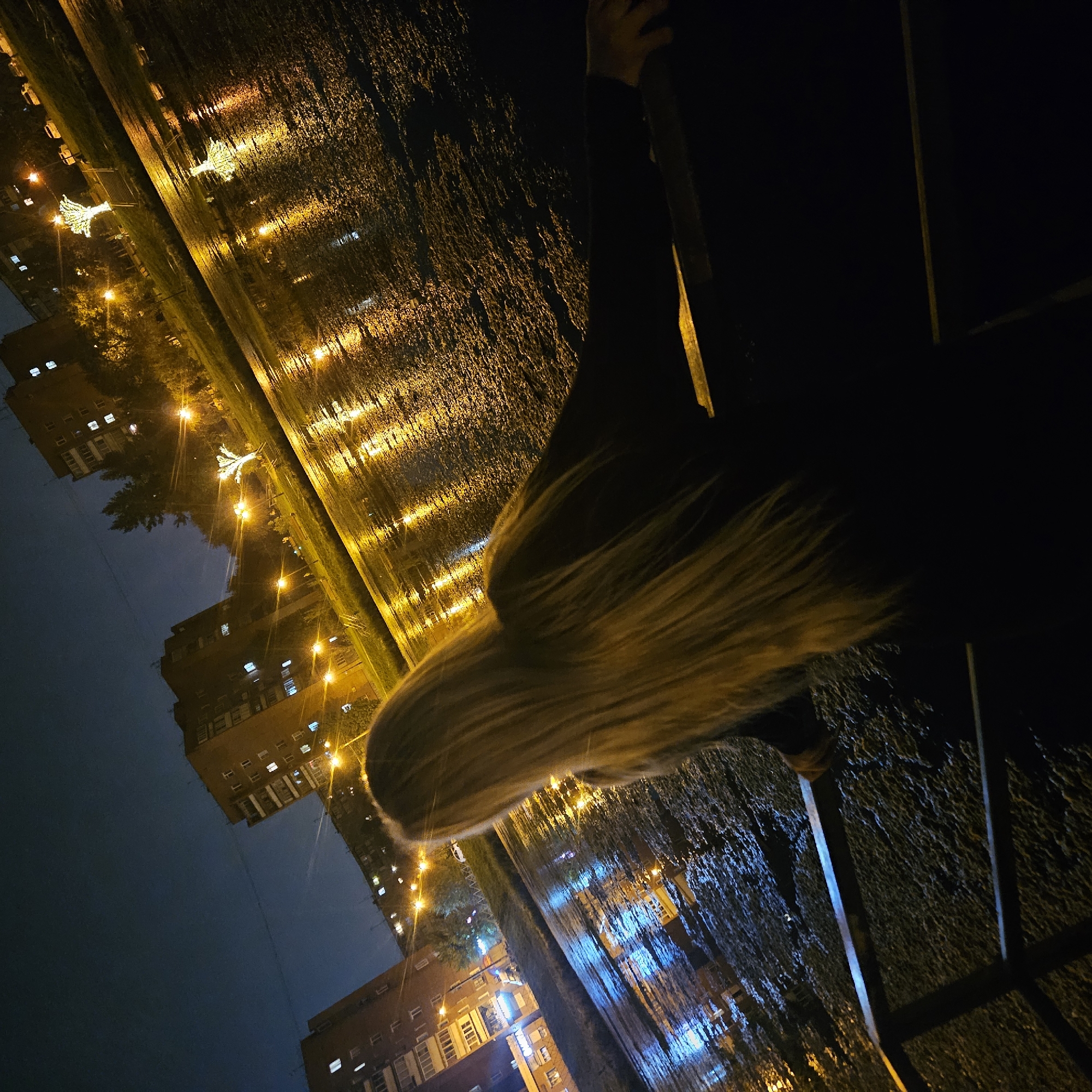The image shows a woman with long blonde hair standing on a street at night. The street is wet, reflecting the lights of buildings and streetlights. In the background, there are apartment buildings and trees. The scene appears to be a city street, possibly a promenade or embankment given the wet surface and reflections. The architectural style of the buildings suggests a post-Soviet era setting. The lighting indicates it is nighttime, and the wet surface implies recent rain or a damp environment. The woman is facing away from the camera, looking out at the scene. The overall atmosphere is somewhat melancholic and reflective, typical of urban night scenes with wet conditions.