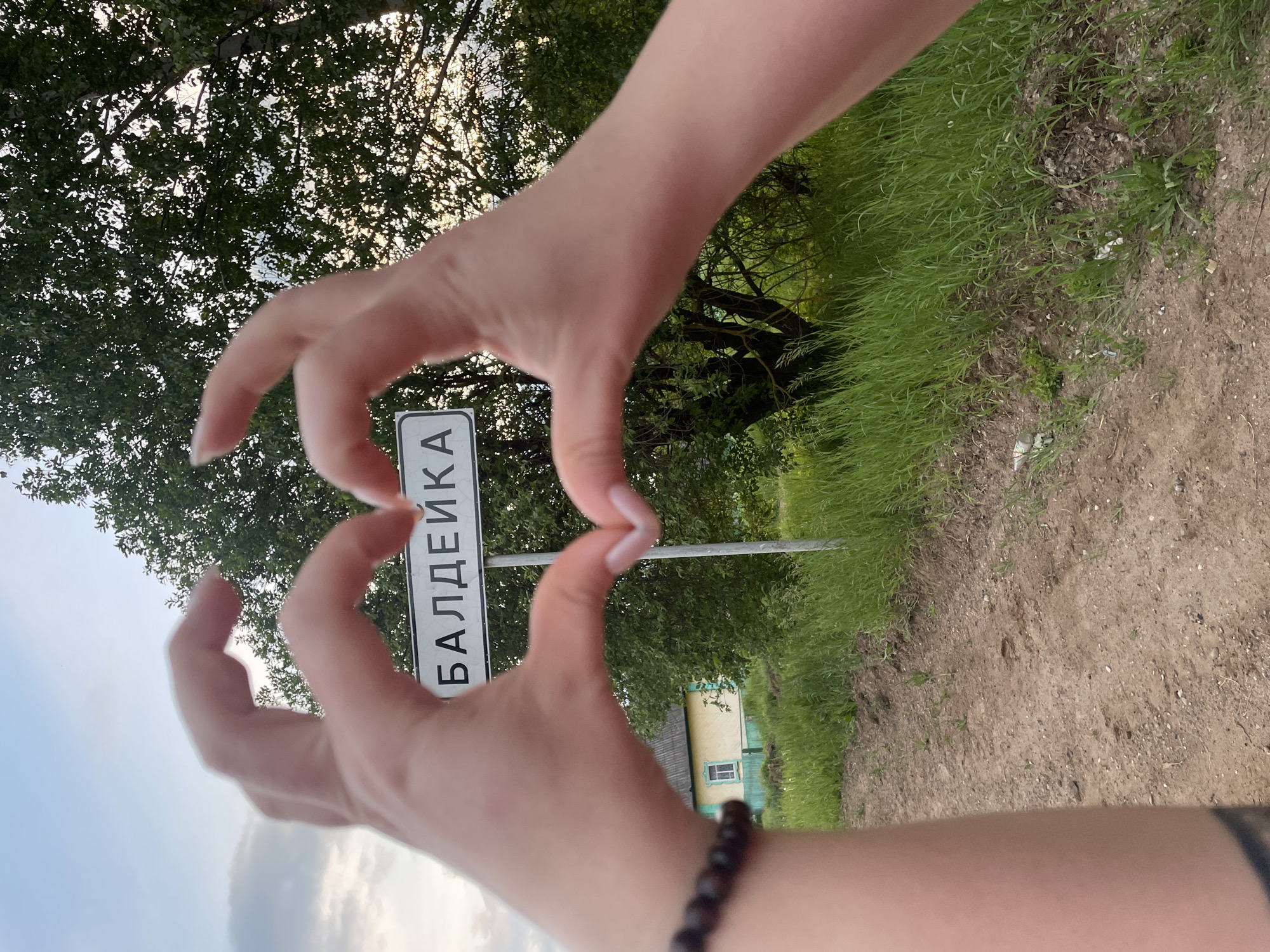 A photograph taken at ground level features a sign indicating the location of Балдейка, Russia. Two hands, framing a heart shape, are positioned in the foreground, partially obscuring the sign. The background comprises greenery and a building with a yellow exterior and blue window trim. The ground is dirt. The left arm has a black bracelet and a tattoo.