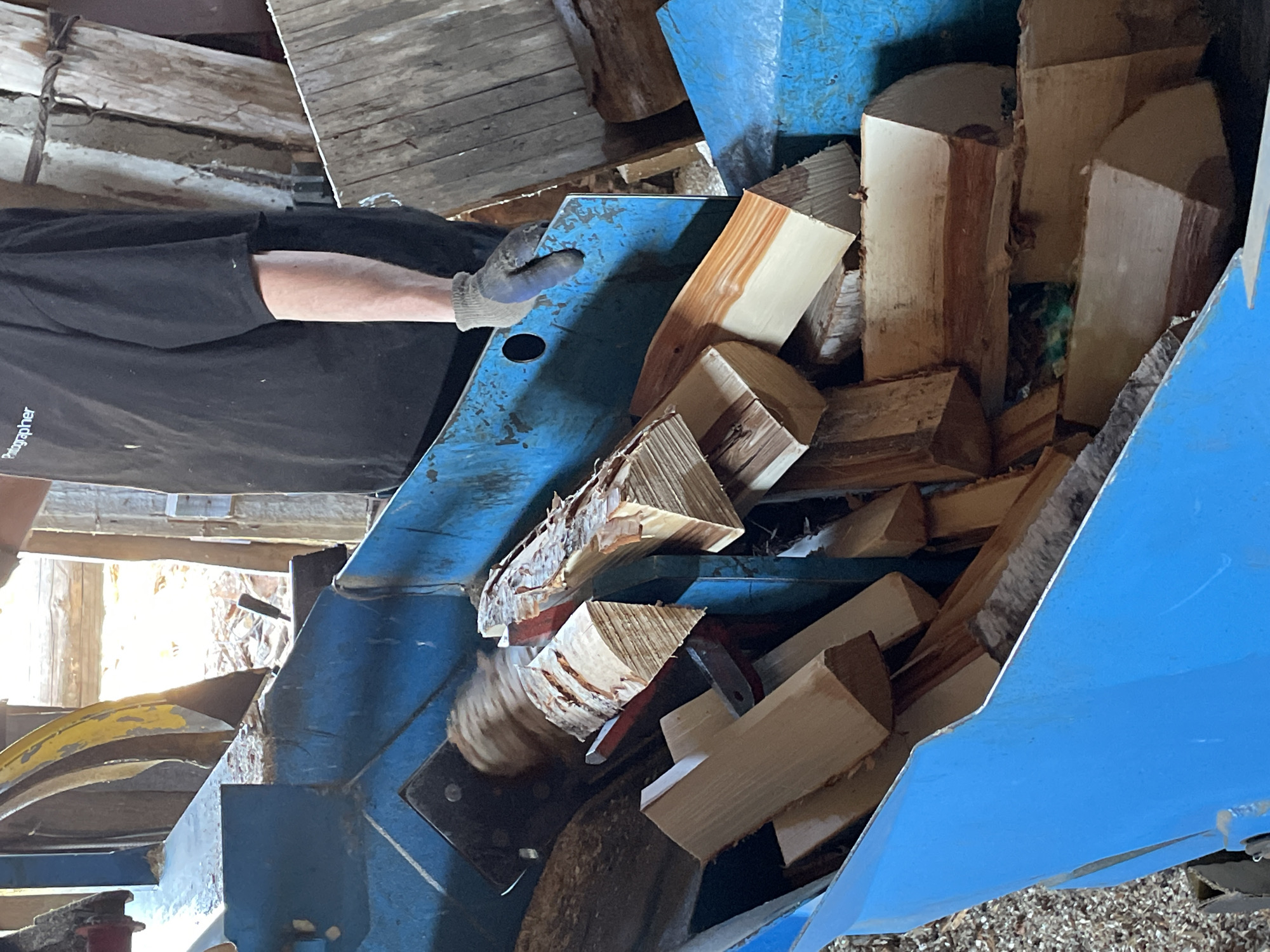 The image depicts a scene inside a wooden structure with a blue, industrial-looking machine filled with cut logs. A person wearing a black shirt and gray glove is positioned to the left of the machine, appearing to interact with it or the logs within. The setting seems to be a work area, possibly a lumber mill or a wood processing facility. The location is specified as Van'ki, Russia, though there is no visual information within the image to confirm this.