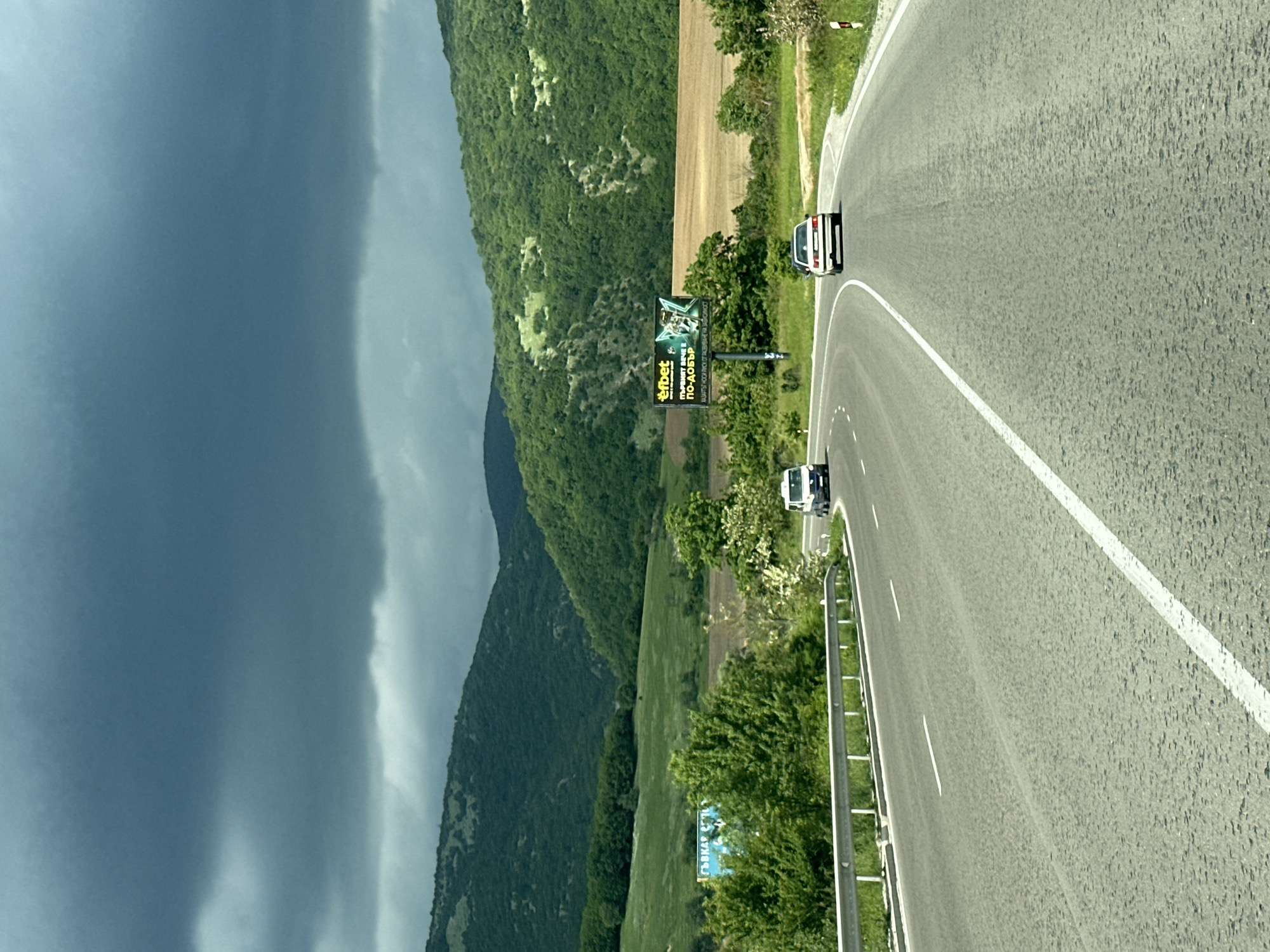 The image shows a winding road in Prolaz, Bulgaria. The road curves through a green landscape of rolling hills and mountains under a stormy sky. Two cars are traveling on the road. Billboards line the roadside. One billboard is branded with "Efbet", another with "Съдийска". The sky is ominously dark, suggesting an approaching storm.