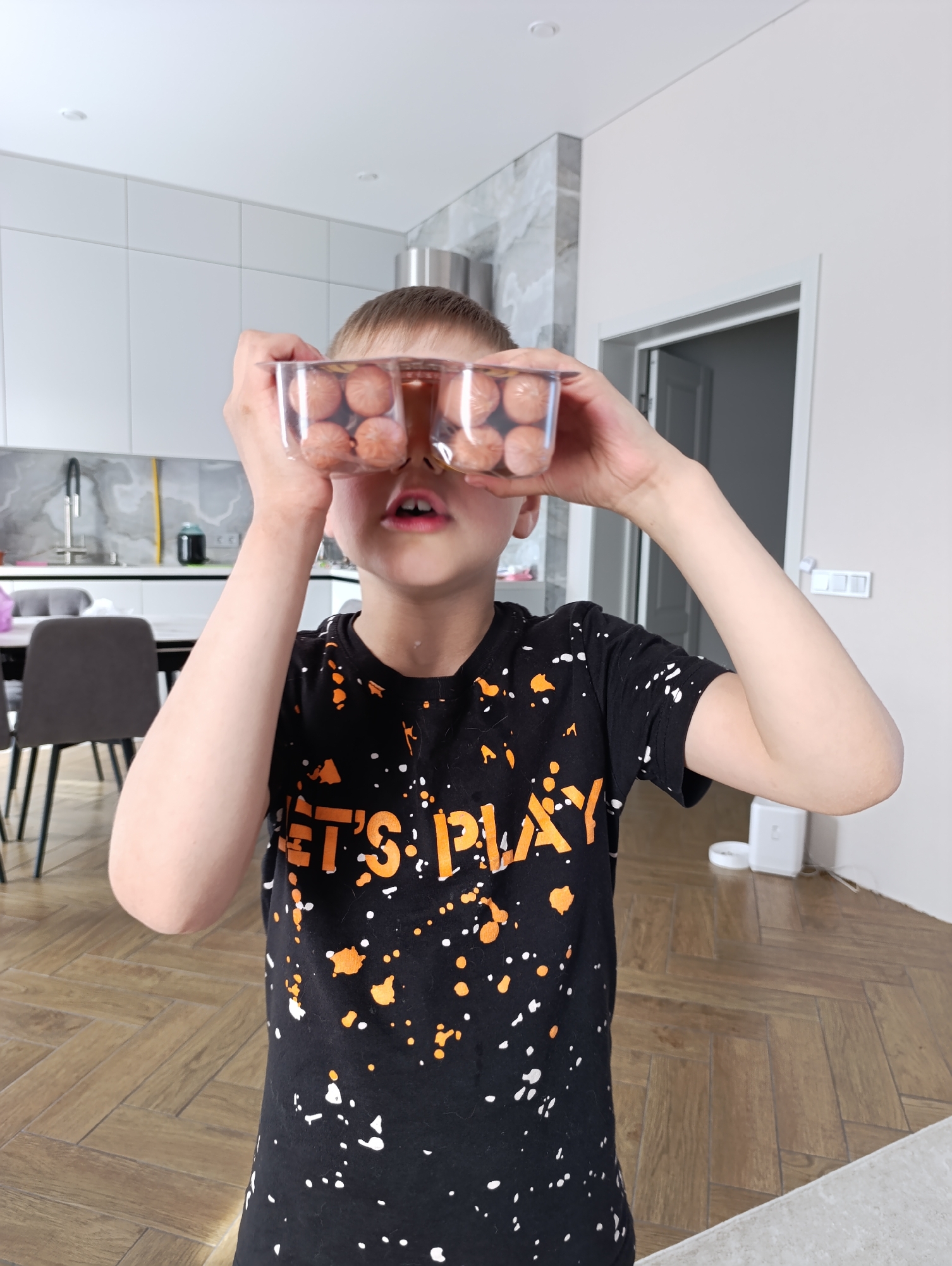 A young boy with light skin and short brown hair stands indoors holding a clear plastic container filled with what appear to be small sausages or cocktail wieners up to his eyes. He is wearing a black t-shirt with the words "LET'S PLAY" in orange lettering and white and orange splatter-like designs on the front. The background includes a kitchen area with white cabinets and a gray marble backsplash, a dining table with chairs, and a doorway leading to another room. The floor is covered with herringbone-patterned wood flooring. The image was captured in a residential location, likely the subject's home, within the city of Chaikovsky, Russia.