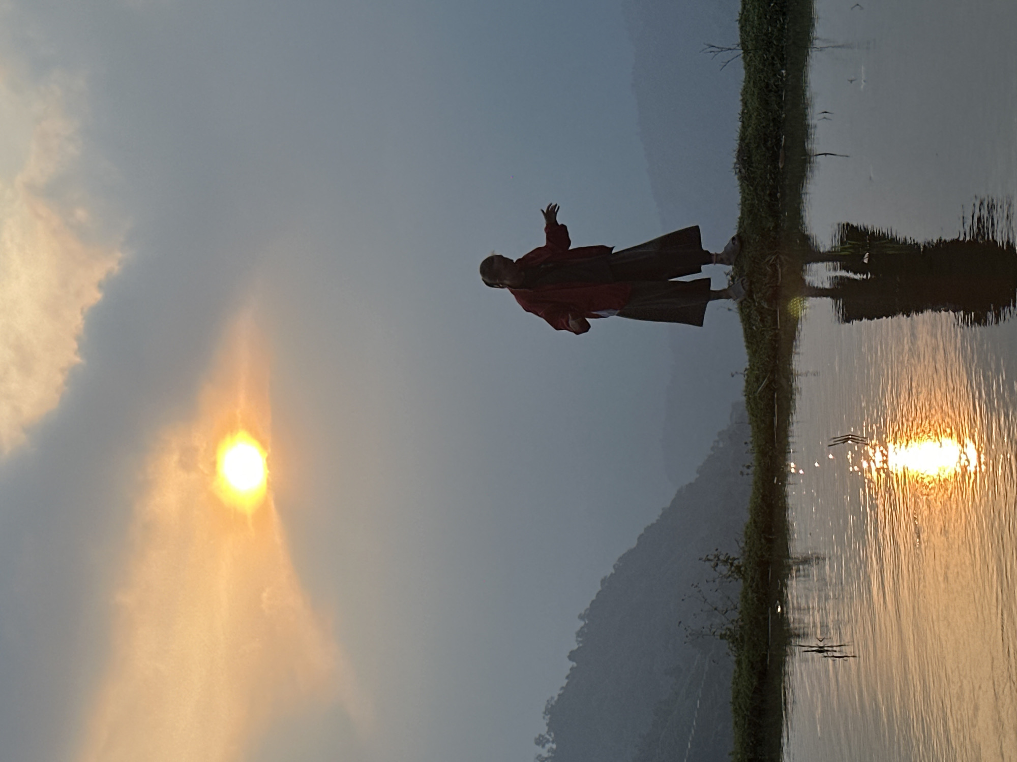 An outdoor landscape photograph, oriented correctly, captures a serene golden hour scene in Nậm Dịch Commune, Vietnam. A lone figure, likely a monk or a person in traditional attire, stands in silhouette near a calm body of water, possibly a flooded rice paddy or a tranquil lake. The figure wears dark pants and a red, flowing top or robe, with one arm raised in a gesturing or contemplative pose. The figure's reflection is clearly visible in the still water.
The sun, a bright golden orb, is positioned on the left side of the frame, partially obscured by soft, hazy clouds. Its warm light casts a brilliant, elongated reflection across the water, creating a shimmering path of gold. The surface of the water perfectly mirrors the figure, the sun's reflection, and the distant, muted silhouette of mountains that frame the background. A thin strip of lush green vegetation lines the edge of the water in the foreground.
The sky is a soft gradient of light blue and grey, with hints of orange and yellow near the sun. The overall atmosphere is peaceful and contemplative, highlighting the natural beauty of the Vietnamese landscape at either sunrise or sunset. No discernible text is visible in the image.