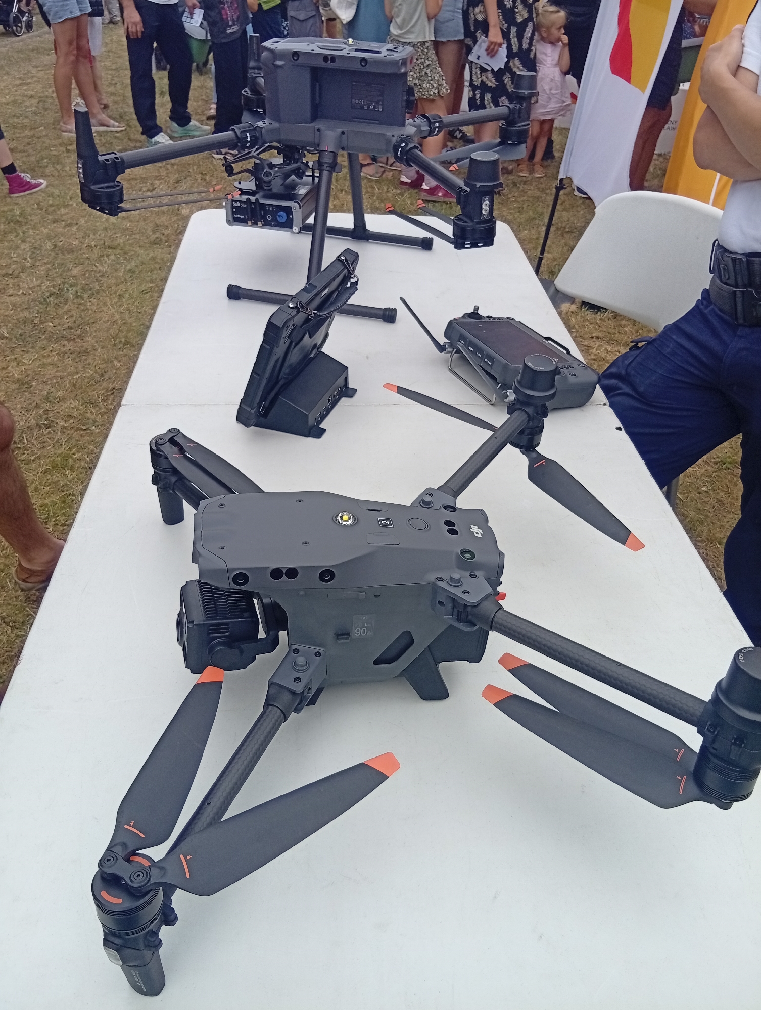 The image shows two large drones and a controller placed on a white table, likely at an outdoor event. The drones are gray with black propellers, some tipped with orange. The larger drone in the foreground is a DJI Matrice series, with its camera gimbal visible. Behind it, another similar drone is partially visible, with its battery pack and other accessories attached. A drone controller is positioned to the right of the larger drone. In the background, several people are standing, some of whom appear to be onlookers or participants in the event. The ground is covered in dry grass, suggesting an outdoor, possibly park-like setting. The overall impression is of a technology demonstration or exhibition. The image was taken in Wrocław, Poland, though the specific location within the city is not evident.