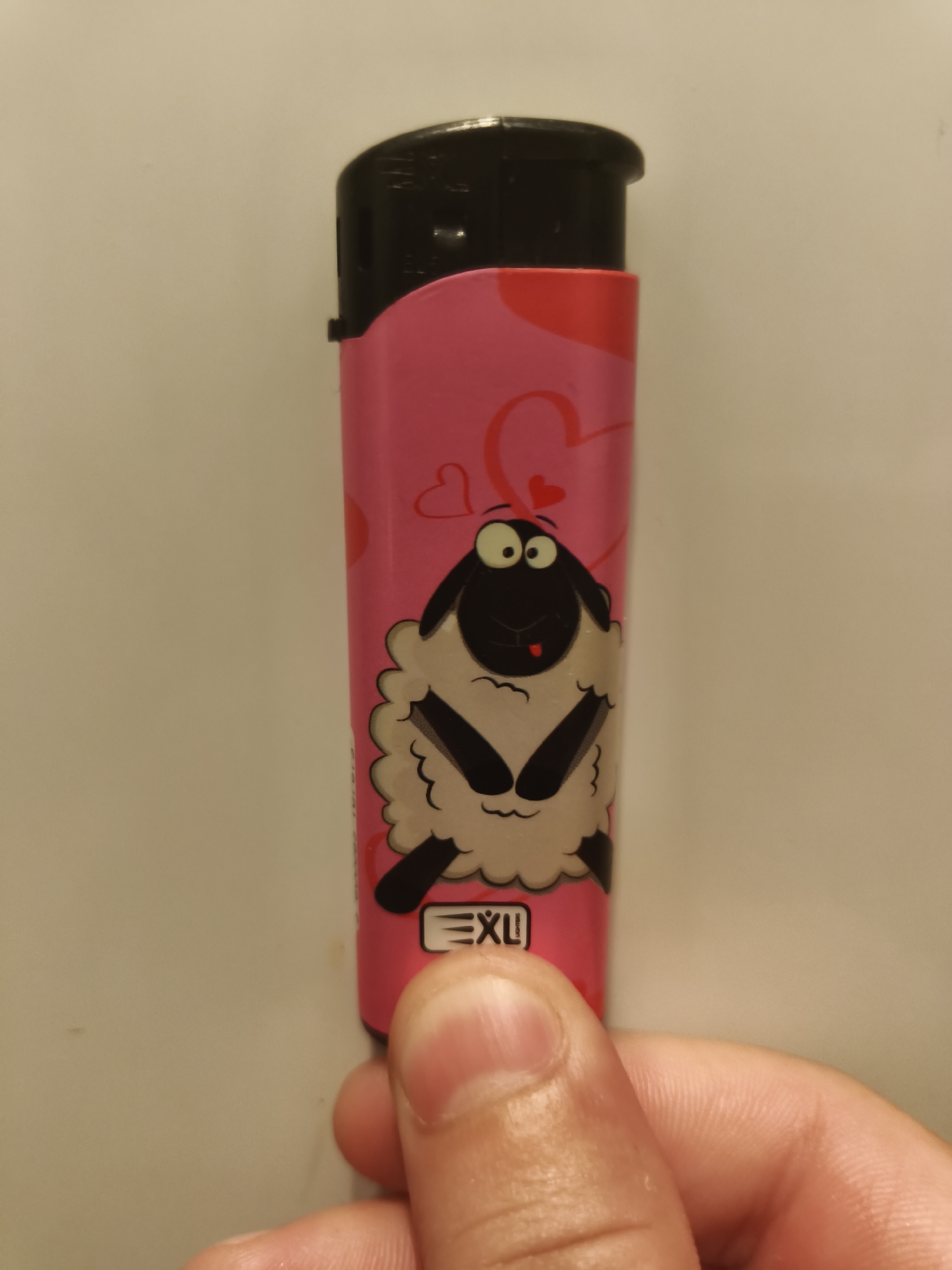 A close-up, eye-level shot captures a person's hand holding a pink lighter against a plain, light-colored background. The hand, with visible fingers and thumb, holds the lighter vertically, with the thumb positioned near the bottom of the lighter.
The lighter itself is predominantly pink, featuring a whimsical design. A cartoon sheep with a white fluffy body, black face, large white eyes, and black limbs is prominently displayed on the front. The sheep's tongue is playfully sticking out. In the background of the lighter's design, there are several stylized heart shapes in both pink and red. The top portion of the lighter, which houses the ignition mechanism, is black. Near the bottom of the lighter, beneath the sheep character, the brand name "EXU" is clearly visible in white capital letters. There are faint, unreadable markings on the black top part of the lighter.
The lighting is soft and even, suggesting an indoor setting. The image focuses on the lighter and the hand, with the background intentionally blurred or out of focus. This scene is observed in Svilengrad, Bulgaria.