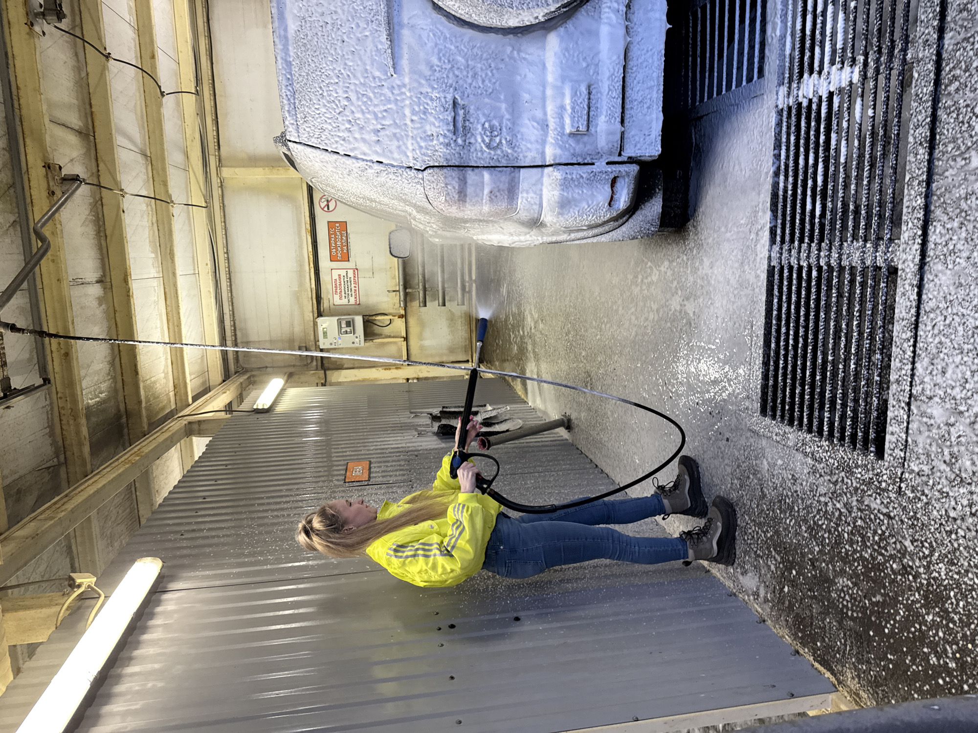 A woman is depicted inside a self-service car wash bay in Chaikovsky, Russia, actively washing a light-colored SUV. She stands on the left side of the bay, wearing a bright yellow long-sleeved jacket with three dark stripes on the sleeves, blue jeans, and dark grey high-top boots. Her blonde hair is tied in a high ponytail. She holds a pressure washer with both hands, aiming the nozzle at the rear of the SUV, which is heavily coated in white soap foam. The vehicle rests on a wet, foam-covered concrete floor that features dark metal drainage grates in the foreground. The car wash bay is enclosed by corrugated grey metal walls; the ceiling is concrete with exposed beams and fluorescent strip lighting. On the wall behind the car, two white signs with red Cyrillic text are visible: one details equipment usage rules, advising against frequent trigger pressing, and the other states that vehicle wiping must be performed outdoors. A smaller orange sign with "ПЕНА" (Foam) in black Cyrillic text is visible on the corrugated wall to the left of the woman.