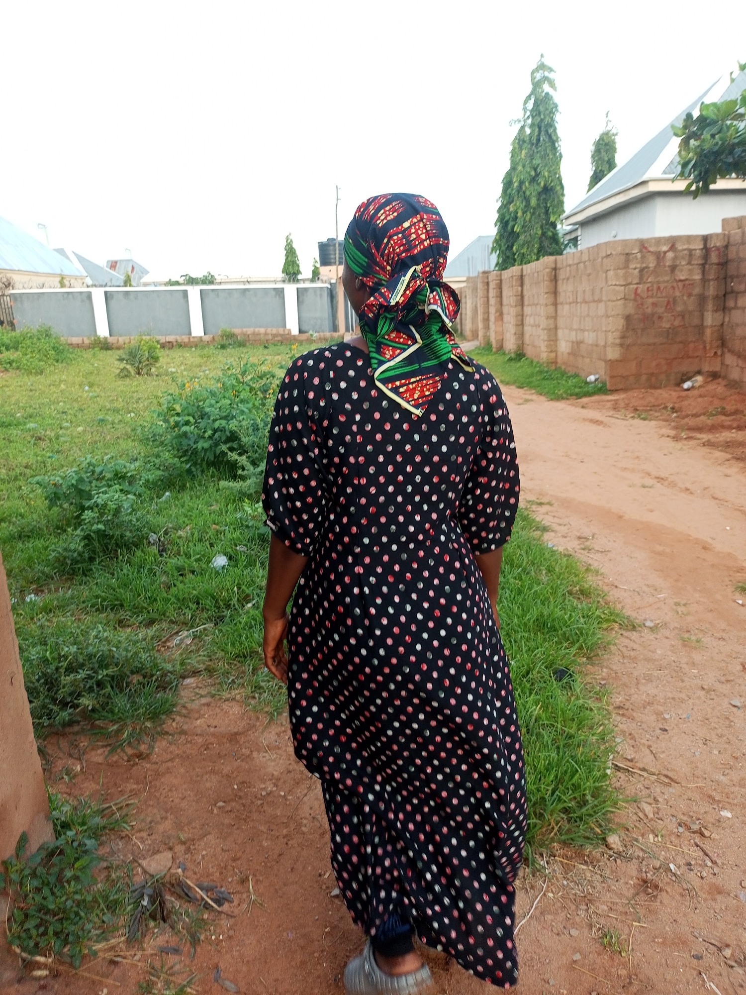 An outdoor, over-the-shoulder shot captures a person from the back, walking on a dirt path in Tempure, Nigeria. The individual wears a black dress with scattered red and white polka dots, and a colorful headscarf with red, green, and yellow geometric patterns. They are wearing gray athletic shoes. To the left of the path, a patch of green grass with scattered weeds and a brown, rough-textured pole are visible. Beyond the grass, a white wall with a gray band runs horizontally across the scene, with buildings and tall, slender green trees behind it. To the right of the path, a low brick wall stretches into the background. The ground is a mix of red dirt and scattered small stones, with some litter present. The sky is overcast and white.