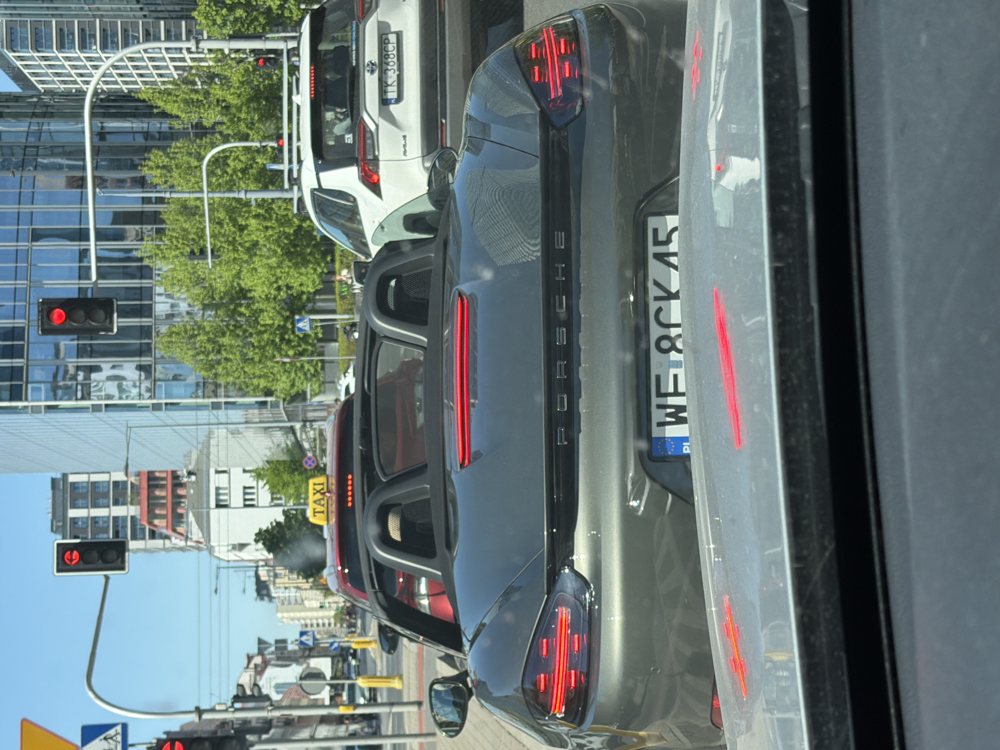 The image shows a view from inside a car, likely in a city, as evidenced by the surrounding architecture and traffic signals. The focus is on the rear of a gray Porsche with a Polish license plate (WE 8CK45). Next to the Porsche is a silver car, partially visible. In front of the Porsche is a white SUV with a license plate (TK 368CP). Further ahead, there is a taxi with a roof-mounted sign. The background features a mix of buildings, including a modern glass building, trees, and traffic lights. The sky is blue, suggesting it is daytime. There's no visible people. The location appears to be a city street with traffic congestion.