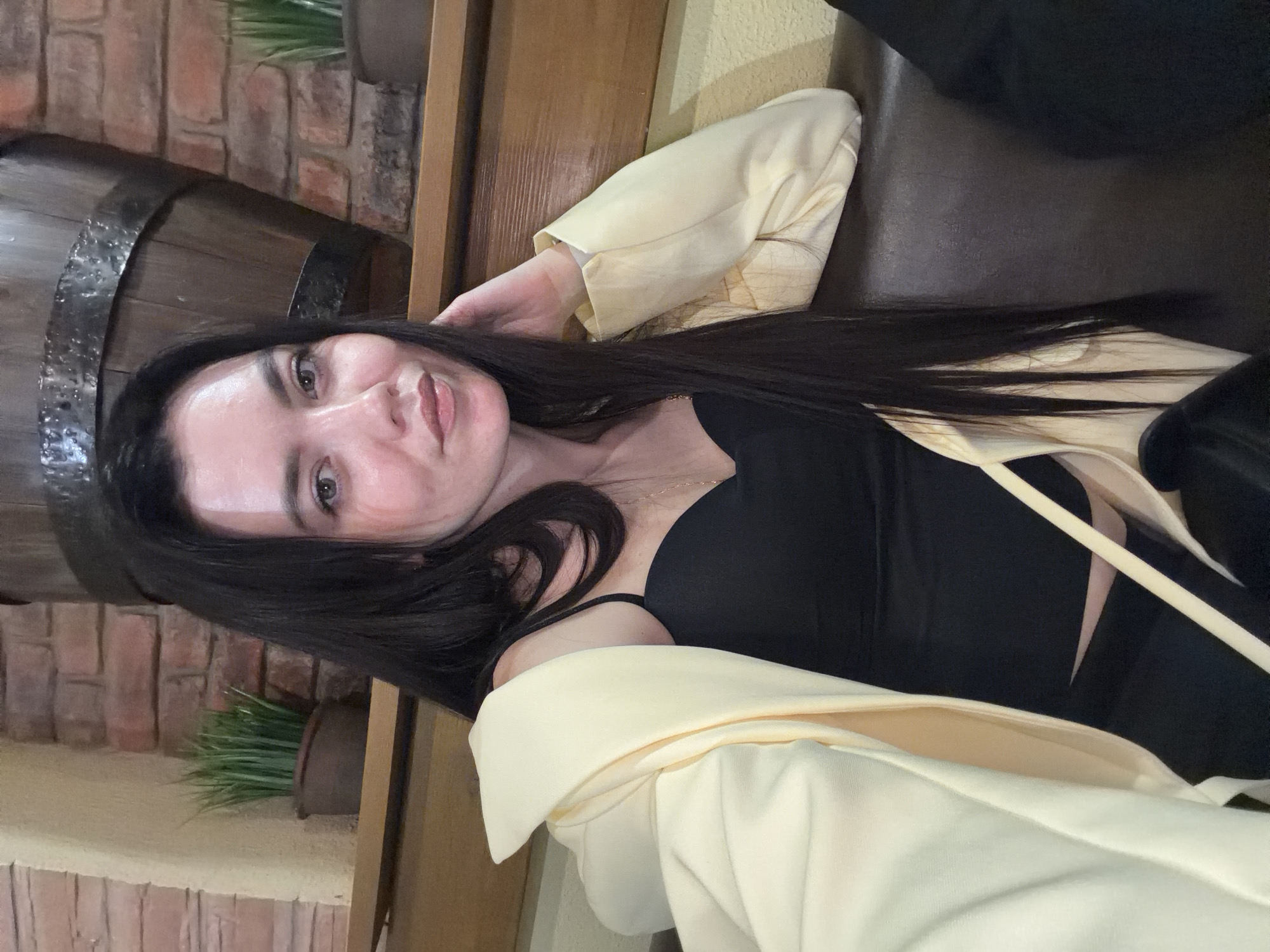 A woman is photographed indoors. She is wearing a black top and a pale yellow blazer. She is sitting on a dark surface, potentially a couch or a bench. In the background, there is a brick wall with decorative items, including a barrel and a plant in a pot. The location is likely a restaurant or bar in Chaikovsky, Russia.