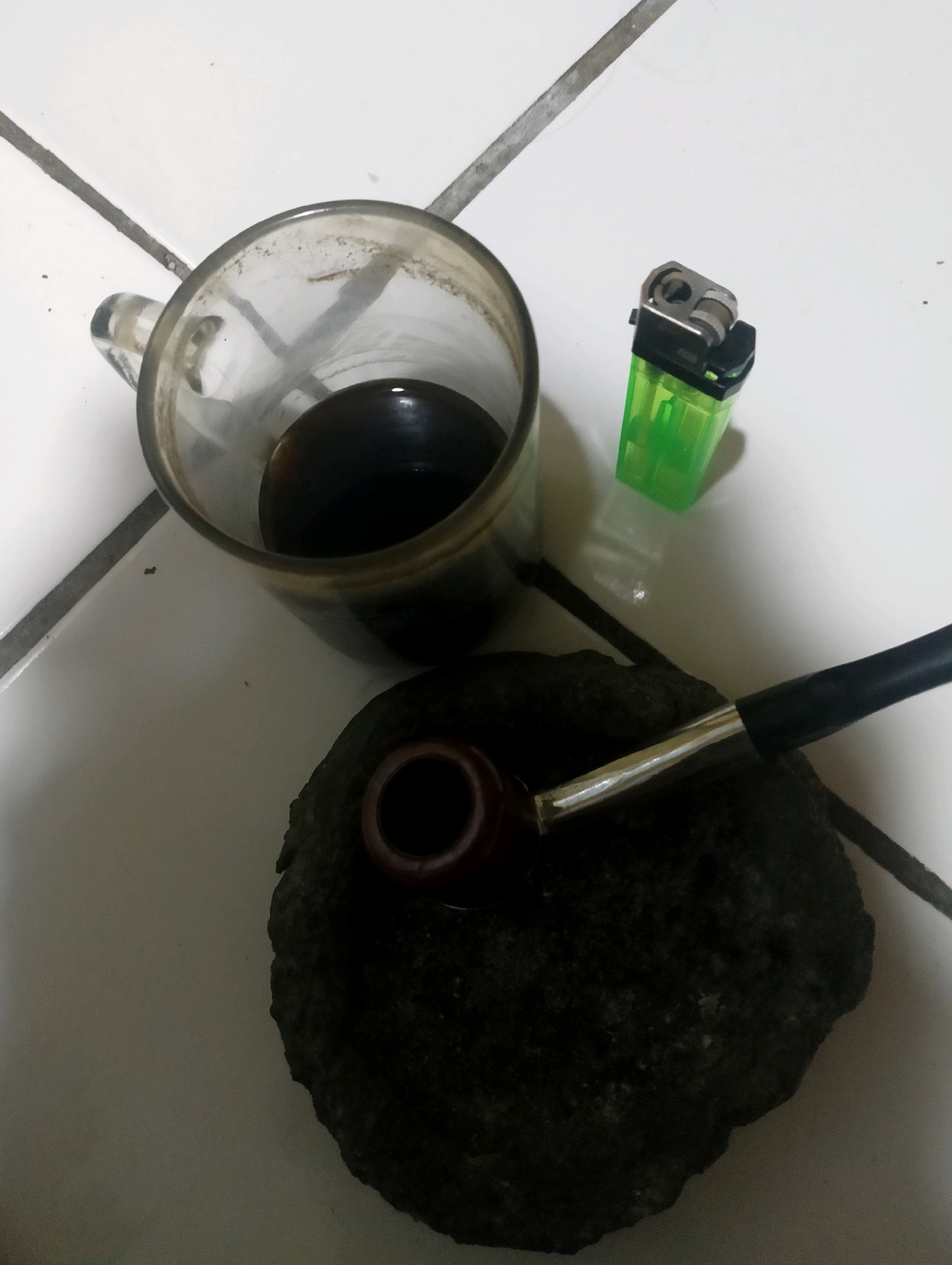 In the city of Ciomas, Indonesia, on a white tiled floor, a scene unfolds. A clear glass mug filled with dark liquid, likely coffee, sits tilted on its side. Beside it, a bright green disposable lighter rests. In the foreground, a dark, irregular stone serves as a base for a tobacco pipe. The pipe, with its polished brown bowl and black stem, is positioned horizontally across the stone, its metallic shank gleaming. The overall impression is one of a personal moment, a pause for reflection or a brief respite.