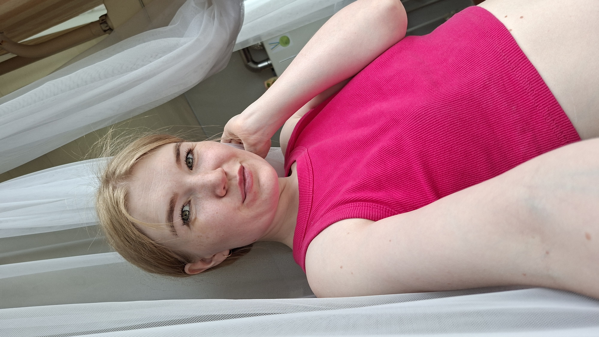 A young woman with blonde hair, green eyes, and a fair complexion with visible freckles is captured in a close-up, angled shot. She is lying down, positioned so her body extends from the lower left to the upper right of the frame, looking directly at the camera with a soft, relaxed expression and a faint smile. She is wearing a bright fuchsia pink, ribbed tank top or crop top.
Her left arm is bent, with her hand resting near her ear and hair, while her right arm is mostly out of frame. Several small moles are visible on her torso just above the waistband of her top.
The setting appears to be indoors, likely a bedroom. White, sheer fabric, possibly curtains or netting from a canopy bed, drapes around her, creating a soft, ethereal background. In the upper left background, a wooden structure, possibly part of a bed frame or furniture, is visible. On a wall behind her, a small green object with unreadable white text or symbols (including what appears to be "60") can be faintly seen. The overall lighting suggests daytime. The context provided indicates the location is Большой Букор, Russia.