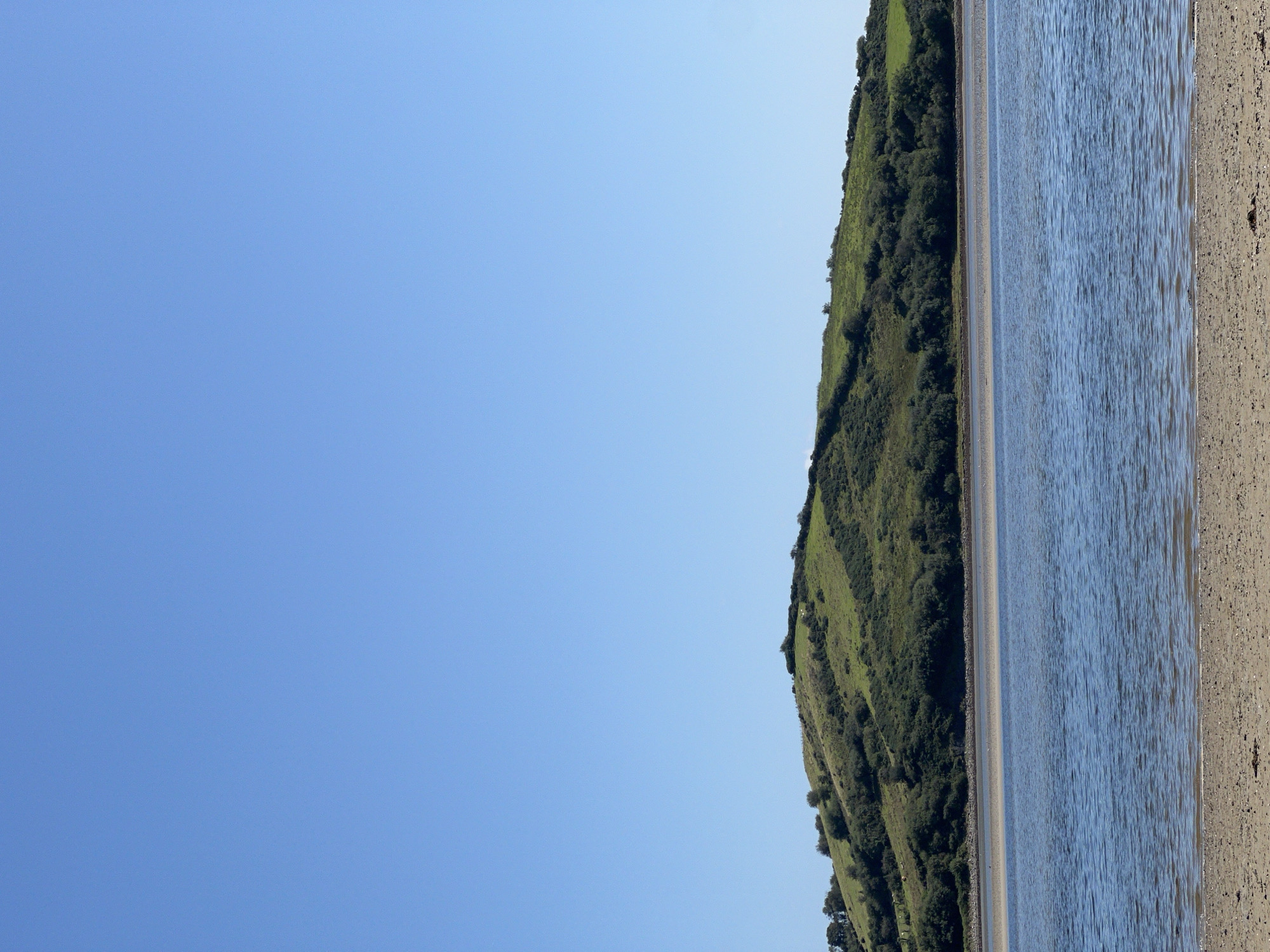 An eye-level, full shot captures a tranquil scene in Tullycullion, Ireland. The upper two-thirds of the image are dominated by a clear, bright blue sky, devoid of clouds. Below the sky, a lush green hillside rises, dotted with scattered trees and bushes. The hillside slopes gently towards a calm body of water that occupies the middle section of the frame. The water is a deep blue, with subtle ripples reflecting the sunlight. In the foreground, a sandy beach meets the water's edge. The sand is light brown and appears speckled with small pebbles. The scene is serene and untouched, conveying a sense of natural beauty and peacefulness.