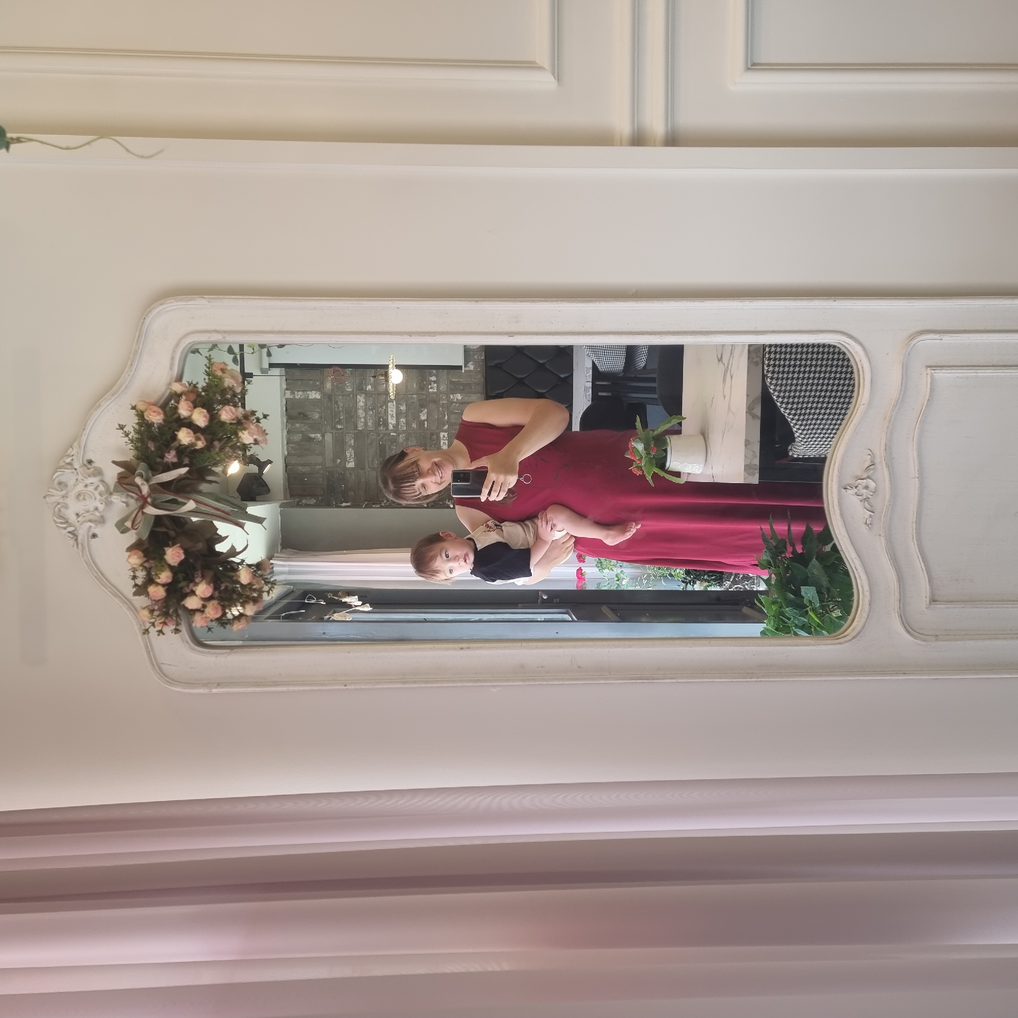 A woman in a long red dress holds a young child in her arms as she takes a selfie in an ornate, distressed white mirror. The child is wearing a black and white shirt. The mirror is decorated with a floral garland and ribbon at the top. The reflection in the mirror shows a brick wall behind the woman and child, a table with a marble top, and chairs with a black and white houndstooth pattern. In the foreground of the reflection, there is a potted plant. To the left of the mirror is a sheer pink curtain. The wall to the right of the mirror is white with decorative paneling. The scene appears to be indoors, possibly in a cafe or restaurant.