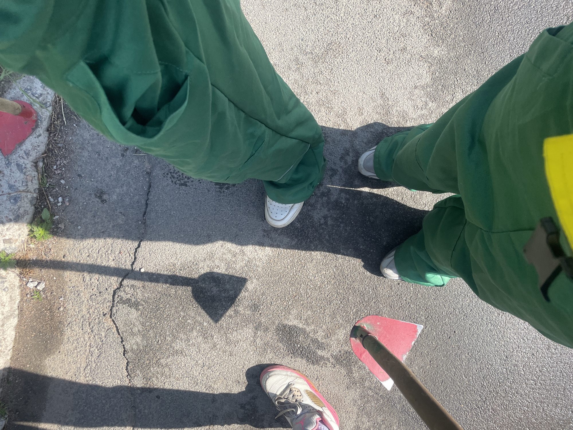 In Saint Petersburg, Russia, a ground-level, top-down perspective captures the lower bodies and feet of at least three individuals wearing matching green work overalls. Two individuals are clearly visible, their legs and feet on an asphalt surface. One pair of feet wears white sneakers, while another wears grey and white sneakers. A third pair of feet, wearing pink and white athletic shoes, is partially visible on the left. The ground is cracked asphalt with some patches of dirt and sparse green vegetation. Shadows of what appear to be tools, possibly shovels or spades, are cast on the ground from an unseen light source, suggesting outdoor work. One red-bladed tool with a wooden handle is positioned in the lower left quadrant of the frame, and another tool with a red head is visible at the top of the frame. The overall scene depicts a moment of rest or preparation for work.