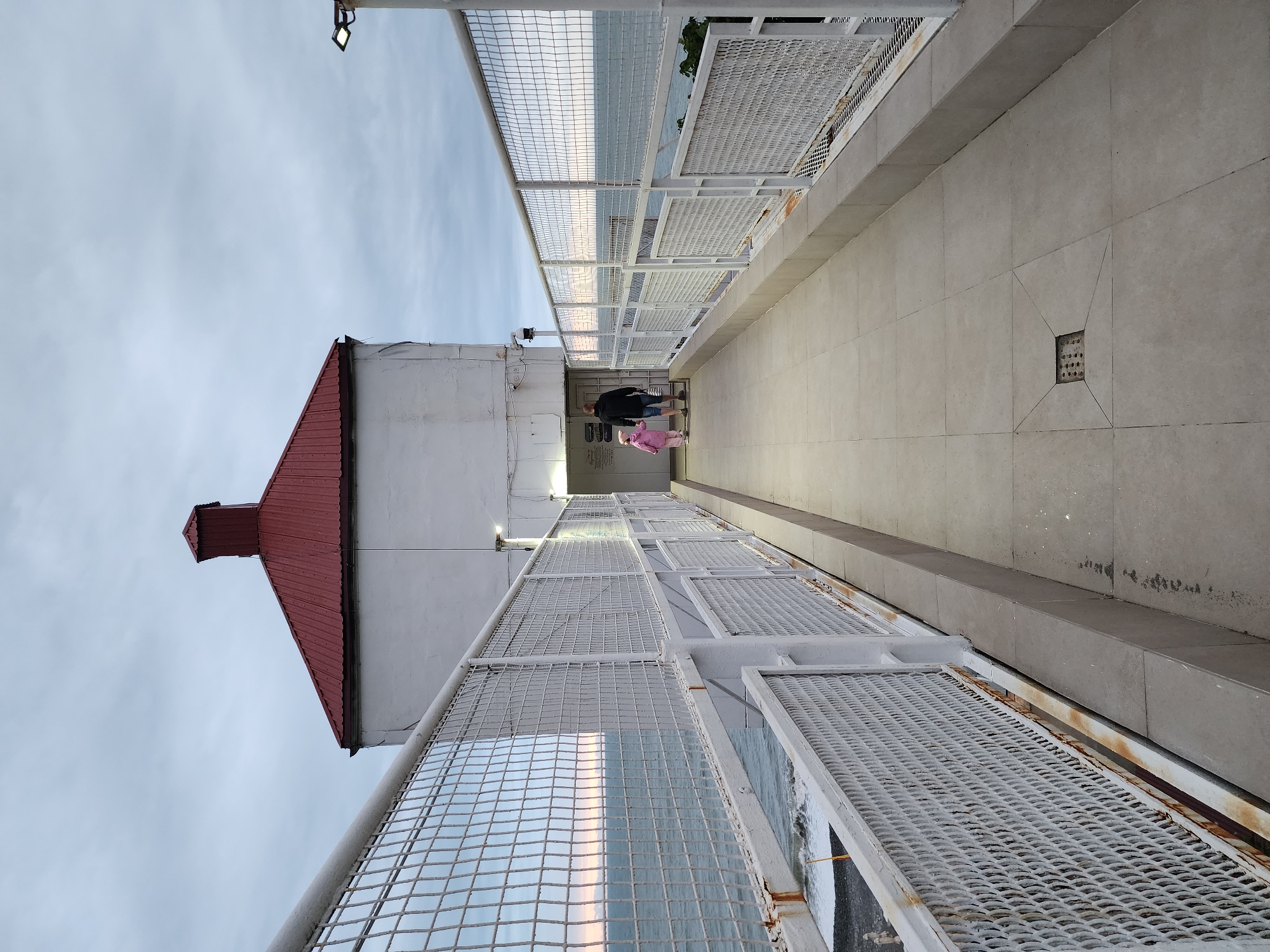 The image shows a walkway in Sochi, Russia, featuring a white metal lattice fence on both sides. In the distance, two figures, one wearing a dark jacket and the other in pink, are walking along the walkway. To the left, a building with a red roof is visible. The sky is overcast. The walkway surface appears to be made of concrete tiles, and there's a square opening in the walkway surface on the right side. There's a lamp post with a light fixture hanging above. Some rust is noticeable on the metal lattice fence.