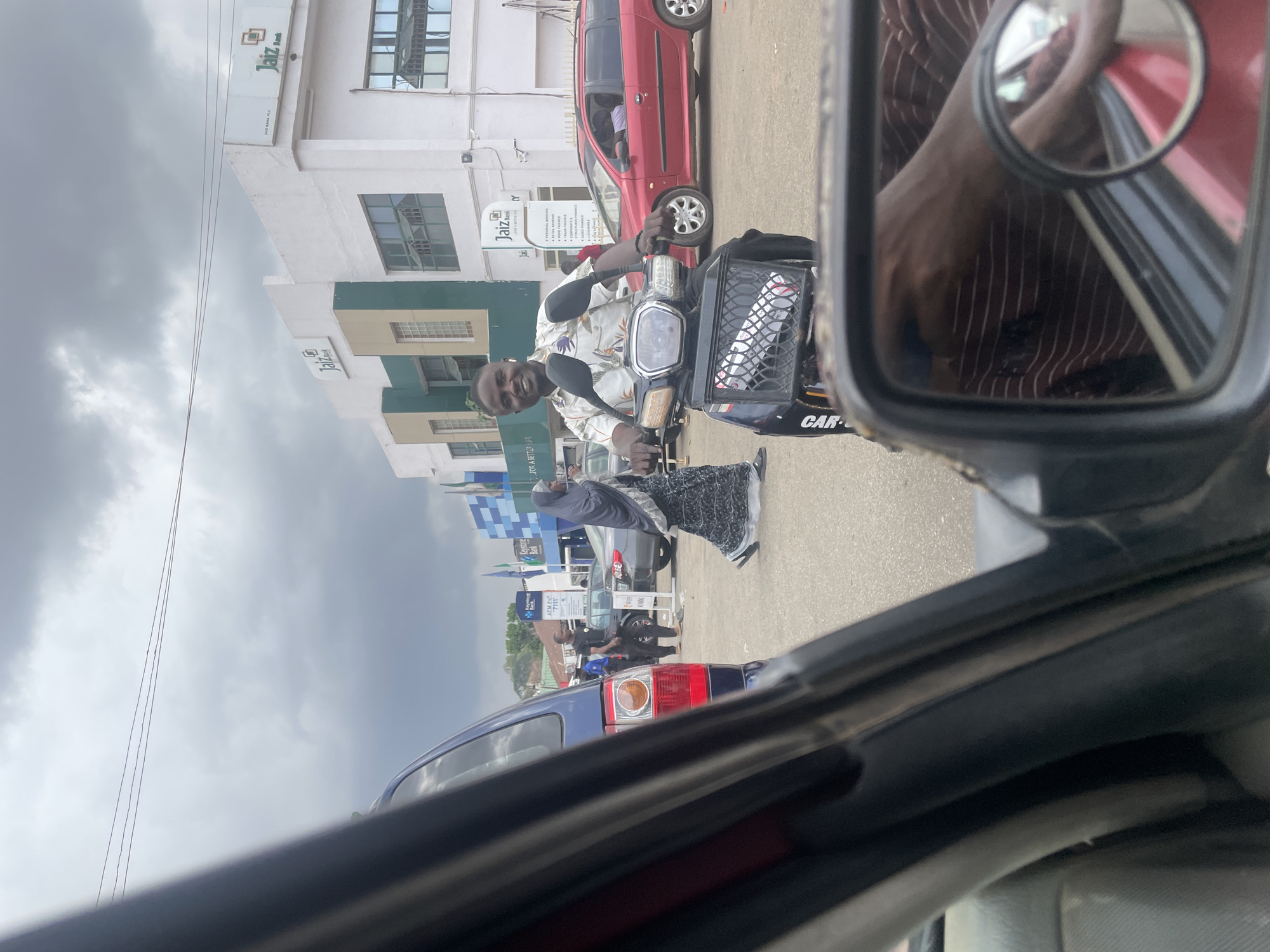 The image, taken from inside a vehicle, captures a street scene in Jos, Nigeria, as evidenced by the presence of "Jaiz Bank" signs in the background. A man on a black motorcycle is positioned in the foreground, facing the camera. He is wearing a patterned shirt and has a wire basket attached to the front of his motorcycle. A red car is partially visible behind the motorcyclist. Further down the street, there is a blue van and several other individuals, including a woman wearing a hijab, near an ATM. The sky is overcast. Parts of the vehicle from which the photo was taken are visible, including the side mirror and the window frame.