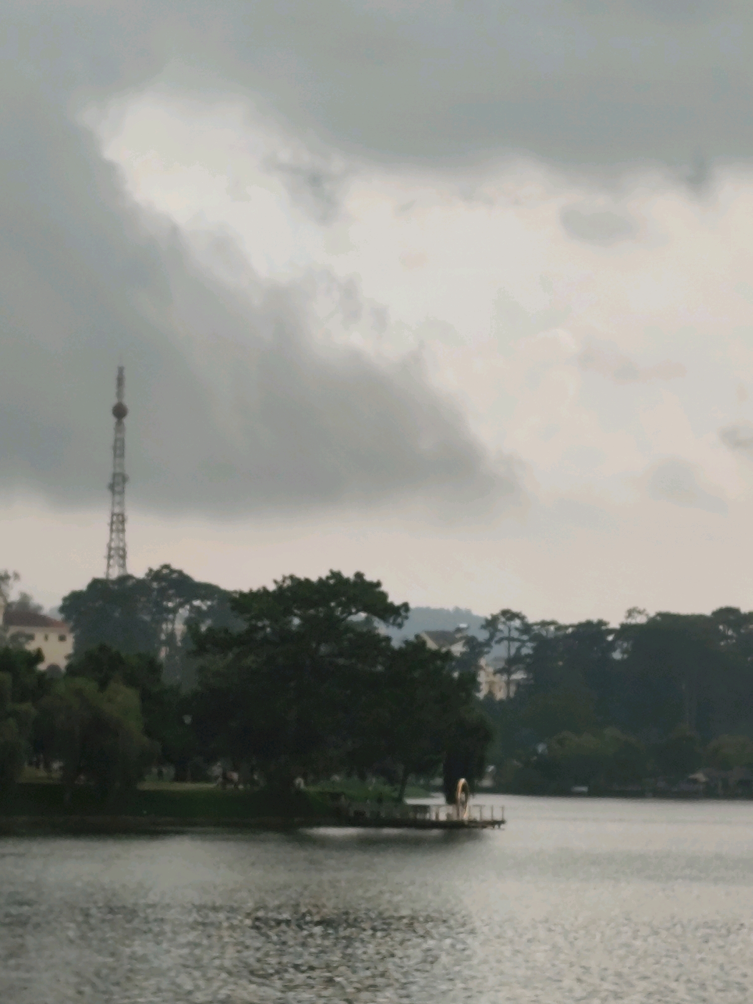 The image depicts Xuan Huong Lake in Da Lat, Vietnam, on a cloudy day. The lake occupies the foreground, with a tranquil surface reflecting the overcast sky. In the middle ground, the shoreline is lined with lush green trees and a grassy embankment. A tall, lattice radio tower is visible on the left side of the image, rising above the trees and silhouetted against the sky. Further along the shore, a small wooden pier extends into the lake. On the pier, a circular, illuminated object, possibly an art installation or decorative feature, is visible. Buildings with light-colored facades are interspersed with the trees in the background, suggesting a built-up area surrounding the lake. The overall atmosphere is serene and somewhat subdued due to the cloudy weather.