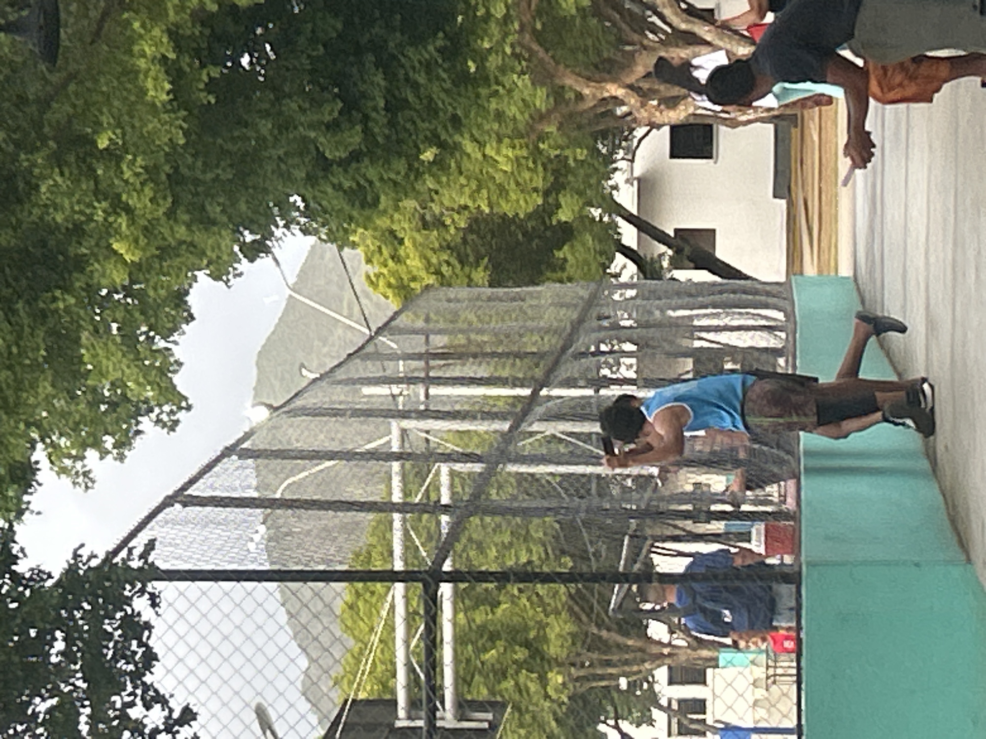 The image captures an outdoor scene at a sports court in Santa Ana, Venezuela. The court is enclosed by a tall chain-link fence. In the foreground, a group of individuals are engaged in activities, with some appearing to be playing a game. One person, wearing a blue tank top and brown shorts, is in motion, possibly mid-game. Another person, in a black shirt and orange pants, is seated and extending their hand, holding a small pink object, to someone out of frame. In the background, a person in a blue backpack stands near a red cooler. The environment includes lush green trees, a white building with dark-framed windows, and a mountainous landscape in the distance under a cloudy sky.