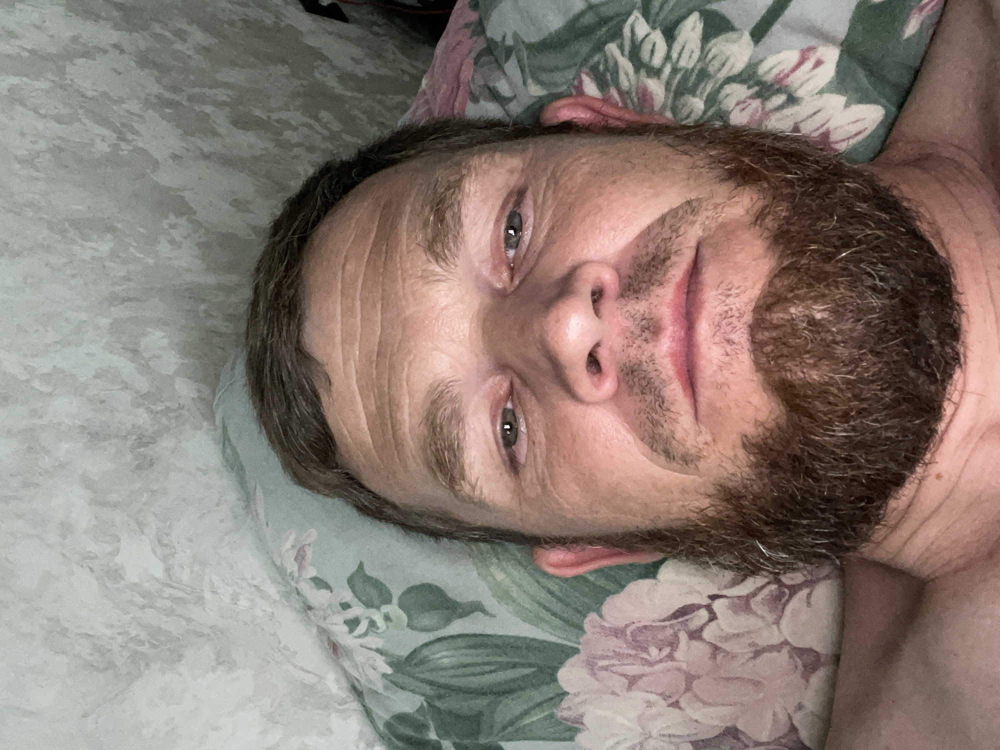 The image shows a close-up, overhead view of a man lying on a floral-patterned pillow. The man has light skin, short brown hair, brown eyes, and a beard. He is looking directly at the camera. The background appears to be a wall with a textured wallpaper pattern. The scene is indoors, likely a bedroom. There is no discernible information in the image that would allow for the specific location within Krasnodar, Russia to be identified.