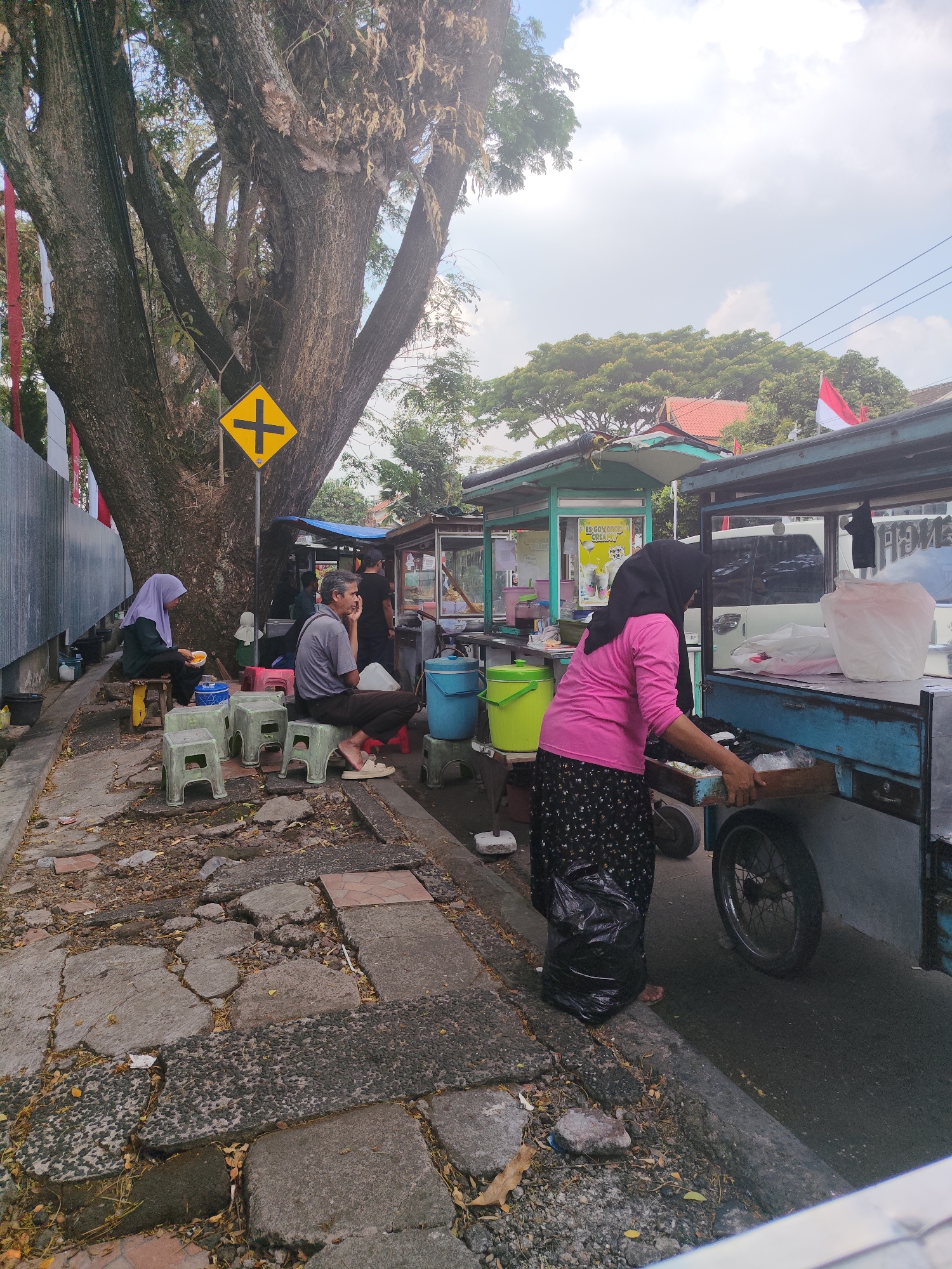 The image depicts an outdoor scene in the city of Garut, Indonesia, likely a roadside vendor area. In the foreground, the cracked and uneven pavement indicates a well-traveled but somewhat neglected urban environment. To the left, a large, mature tree with a thick trunk dominates the frame, its branches extending upwards towards a partly cloudy sky. A yellow traffic sign with a black cross symbol is affixed to a pole near the tree.
Several individuals are present. On the left, a person wearing a purple hijab sits on a small stool, holding a yellow bowl. Nearby, another person in a grey shirt and dark pants sits on a stool, resting their chin on their hand. Further back, other figures are visible near food stalls.
The background features a row of temporary food stalls, identified by their colorful awnings and signage, suggesting a vibrant street food scene. One stall has a sign that appears to advertise "Es Gumbo Cream" and another item priced at "10K." A blue food cart with a wheel is positioned to the right, with a person in a pink shirt and patterned skirt actively attending to it, pulling out a drawer. The cart contains various items, including plastic bags and what appears to be food. Indonesian flags, red and white, are visible on some of the stalls, indicating a sense of local pride or national celebration. The overall impression is one of a bustling, everyday commercial activity taking place along a city street.