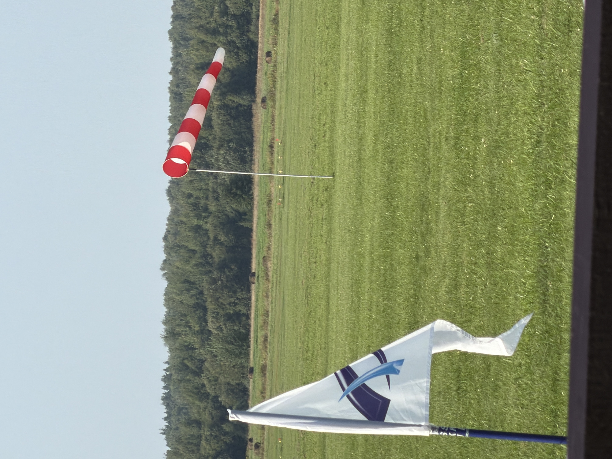 In a grassy field, likely in Korobcheevo, Russia, a white triangular flag with a blue and purple logo is prominently displayed on a blue pole in the foreground. To the right, a windsock with alternating red and white stripes is mounted on a pole, angled upwards, indicating wind. In the background, a dense line of green trees forms the horizon, beneath a clear, pale blue sky. The field is a uniform green, suggesting a well-maintained lawn or pasture. Several dark, circular shapes, possibly hay bales, are scattered across the middle ground of the field. The scene is peaceful and appears to be an outdoor event or recreational area.