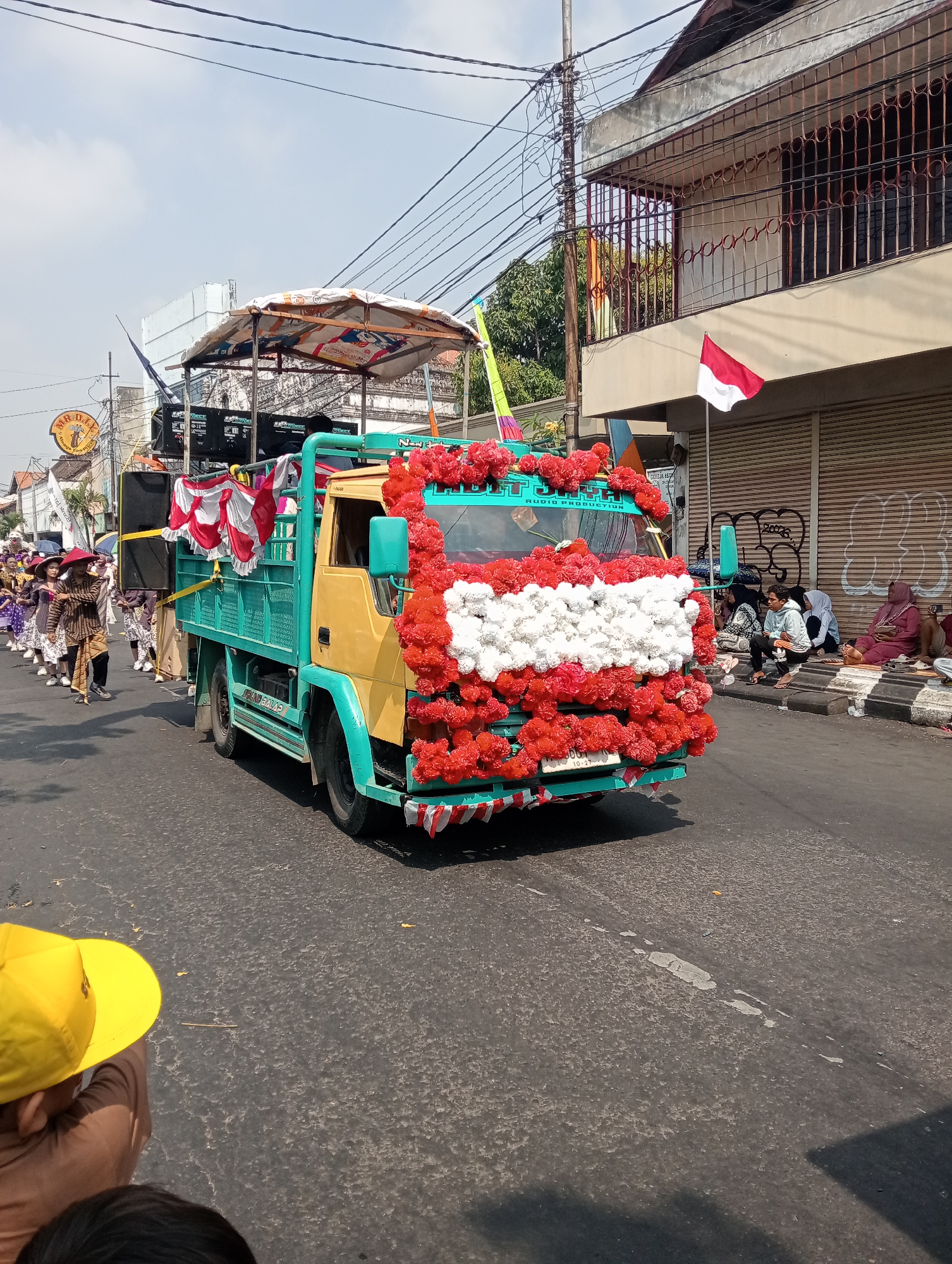 A yellow and teal truck, decorated with red and white flowers, drives down a street in Bintoro, Indonesia. The truck is carrying a sound system and appears to be part of a parade or festival. A group of people in traditional clothing walk alongside the truck. Spectators are seated on the side of the road, watching the procession. The sky is clear and blue, and the sun is shining. The buildings lining the street are typical of Indonesian architecture, with balconies and shops on the ground floor. An Indonesian flag is visible on one of the buildings. The street is paved and in good condition.