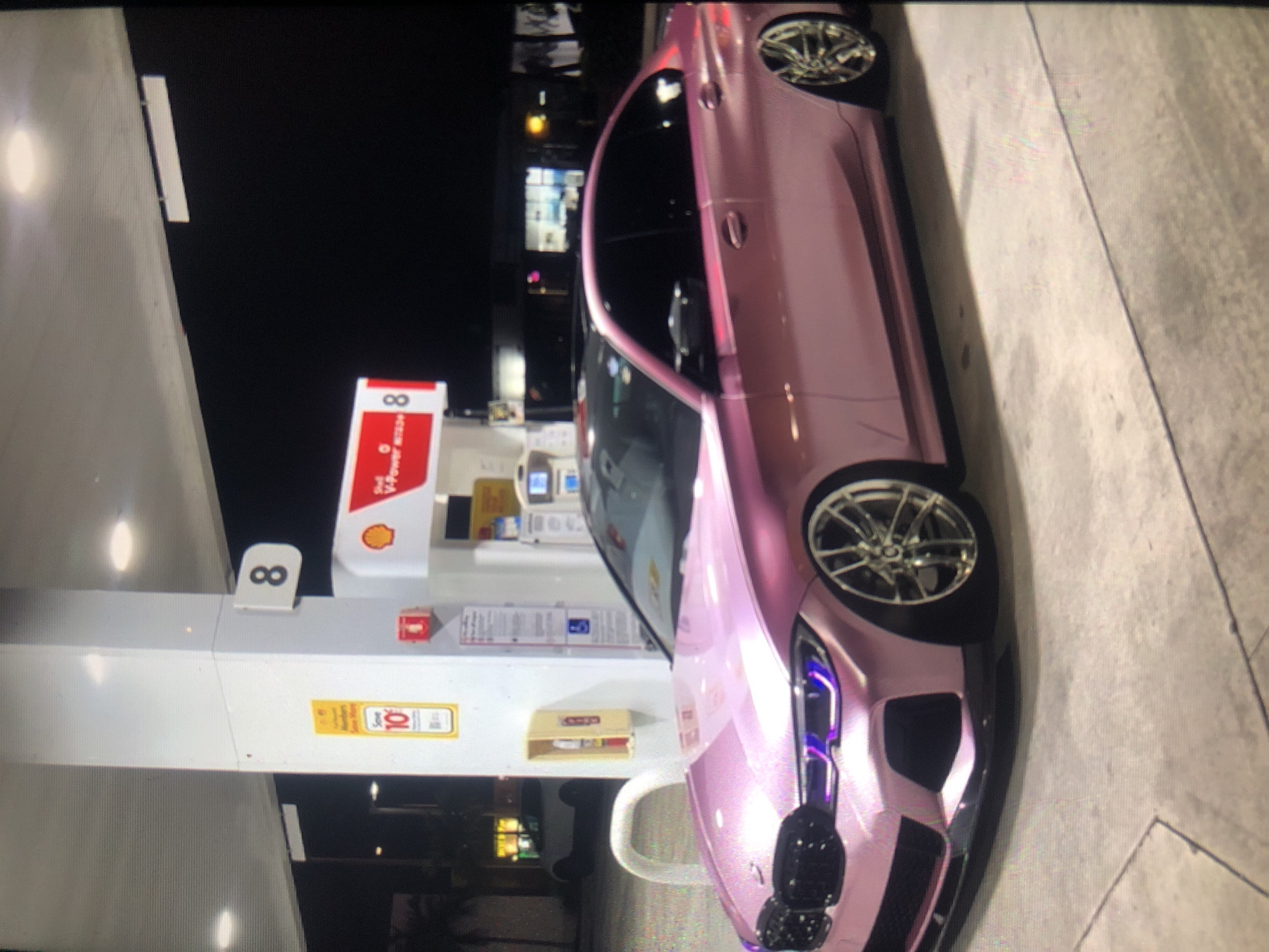 The image depicts a pink sports car parked next to a Shell gas station pump at night. The pump is marked with the number "8." The surrounding environment suggests a well-lit urban area. The image does not show any people. The location within the city of Lhokseumawe, Indonesia is not discernible from the image.