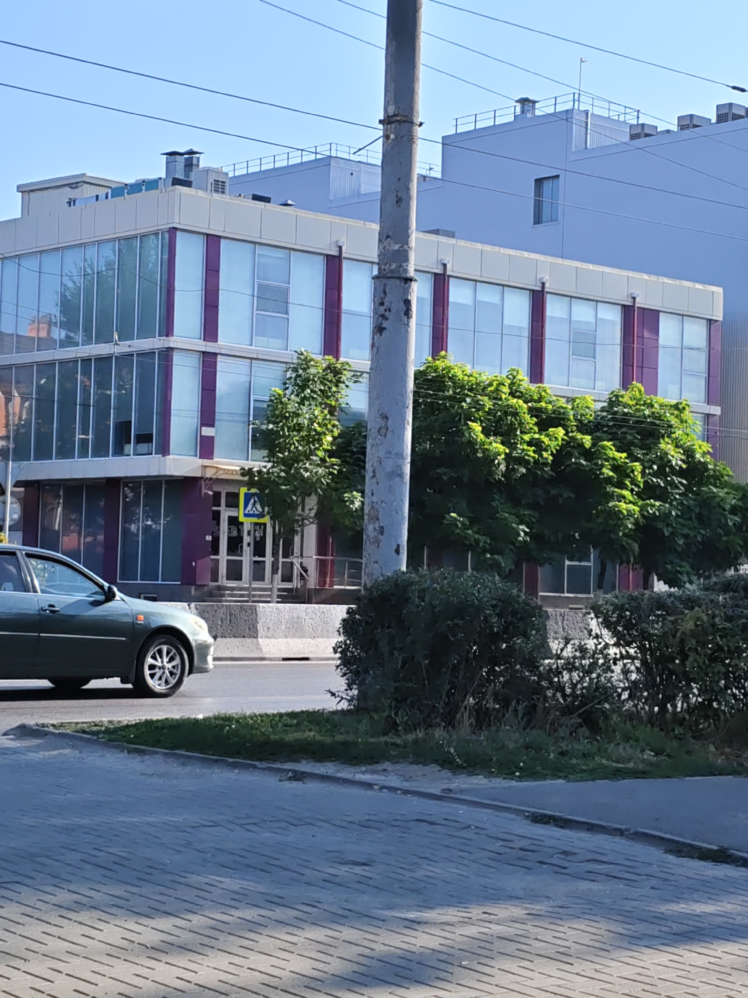 The image captures a street-level view of a modern office building with a glass facade in Rostov-on-Don, Russia. The building features two stories with large windows and distinctive purple vertical accents. In front of the building, there is a pedestrian crossing with a yellow warning sign depicting a person walking. A dark green car is driving on the road in the foreground, to the left of the frame. Lush green trees are visible growing between the building and the road, as well as on the sidewalk. The sky is clear and blue, indicating a sunny day. A tall, weathered utility pole stands prominently in the middle of the frame, obscuring part of the building and trees. In the background, another larger, lighter-colored building is visible, extending upwards and suggesting a more extensive urban landscape. The street is paved with interlocking bricks in the foreground.
