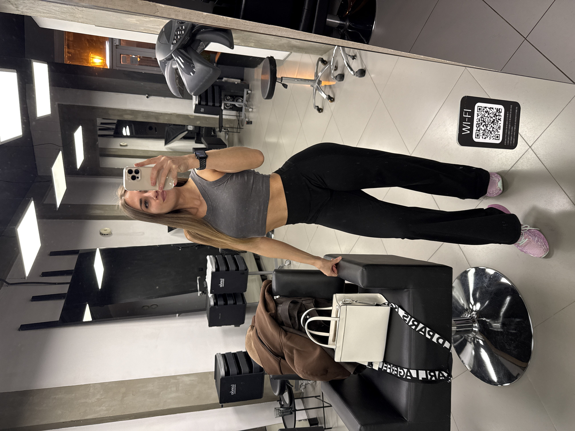 A woman takes a mirror selfie in what appears to be a salon. She wears a gray tank top and black pants. Her blonde hair is long. She holds a white smartphone in her left hand and wears a watch on her left wrist.
The environment includes salon chairs, equipment such as hair dryers, and a white tile floor. A black sign with a QR code for Wi-Fi is visible on the floor. A white bag is resting on a black chair.
The location is potentially Saint Petersburg, Russia, but this is not definitively ascertainable from the image itself.