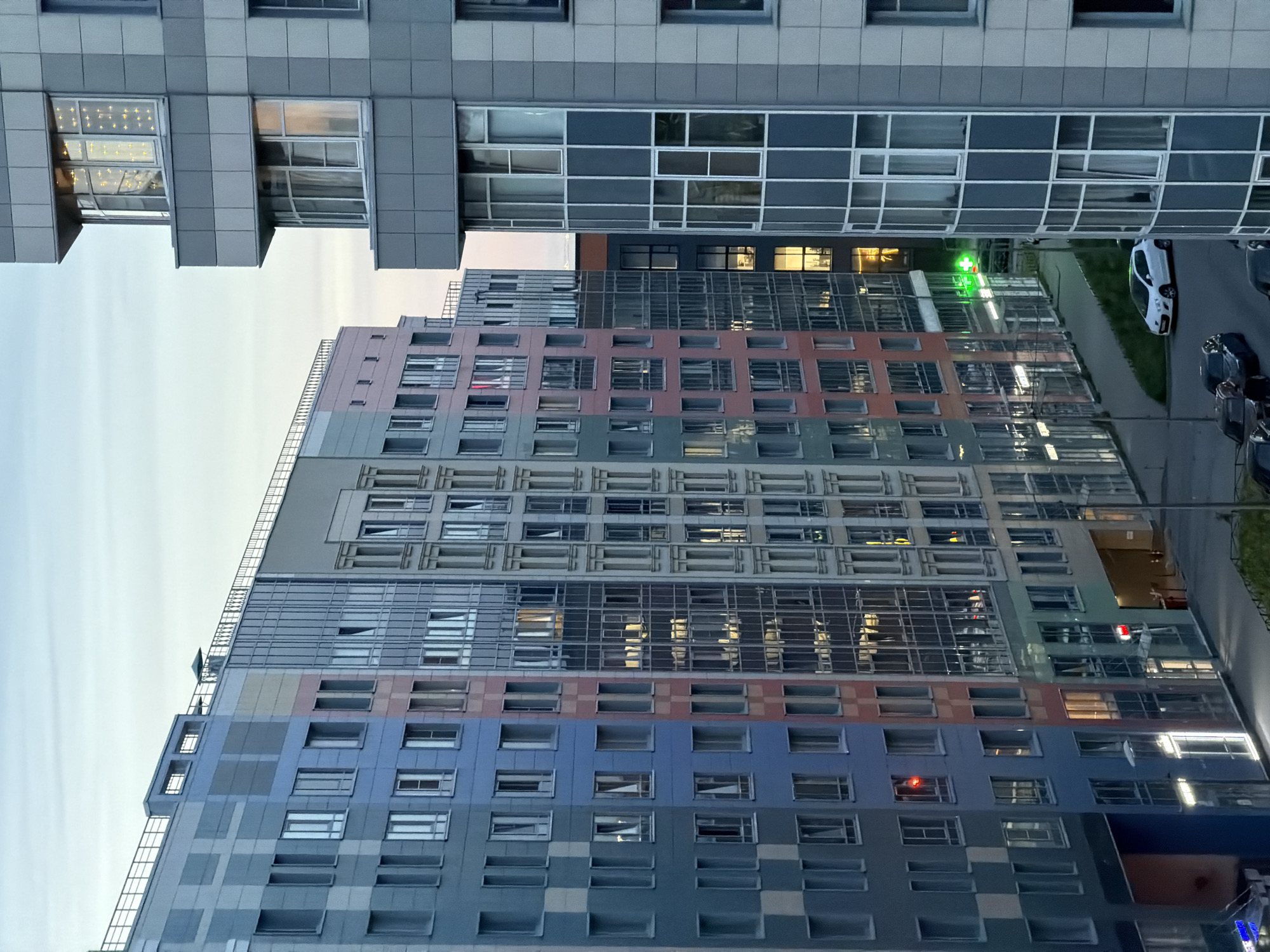 The image displays a modern urban residential environment at dusk or dawn, indicated by the fading light in the sky and illuminated artificial lights. Multiple high-rise buildings dominate the scene.
On the right, a grey multi-story building with distinctive curved bay windows extends vertically, with several windows displaying interior lights. The topmost visible bay window contains numerous small, bright decorative lights.
To the left, a row of multi-colored residential buildings stretches diagonally into the background. These buildings feature facades in varying shades of blue, grey, light pink/salmon, and green, often arranged in block patterns or vertical strips. They possess numerous rectangular windows, many reflecting the environment or showing interior illumination. On one of these buildings, a red traffic light is visible on an upper floor. At ground level, several storefronts are visible; one displays a prominent green cross sign, indicative of a pharmacy, and another has a red rectangular sign.
At street level, a paved road is visible with several parked cars, including a white hatchback and multiple darker sedans or SUVs. A sidewalk runs alongside the buildings. Streetlights are illuminated, casting light on the pavement. No individuals are discernible in the scene.
The architecture and urban planning style, characterized by a mix of colorful, high-density residential buildings with integrated ground-floor commercial spaces, is typical of newer residential developments found in the peripheral districts of Saint Petersburg, Russia, such as Murino or Kudrovo.