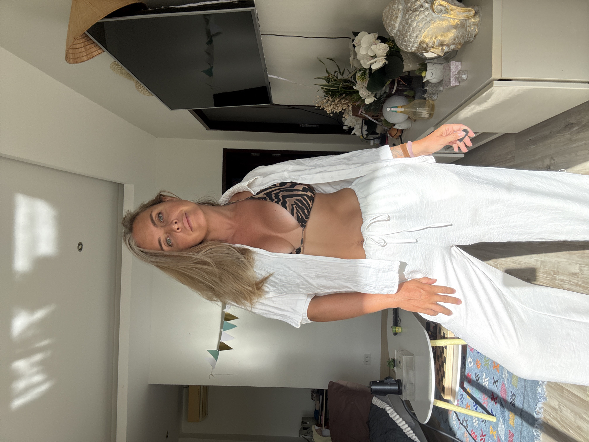 A Caucasian woman with blonde hair is standing in a sunlit room, likely an apartment, in Ko Samui District, Thailand. She is wearing a white, unbuttoned linen shirt over a zebra-print bikini top and loose white linen pants. Her left hand is resting on her hip, and her right hand is holding a small dark object. The room appears to be a living area, with a sofa and a coffee table visible in the background. A television is mounted on the wall, and a decorative Buddha head sits on a cabinet. Sunlight streams through a window, casting shadows on the ceiling and floor. The overall impression is one of a casual, indoor setting on a bright day.