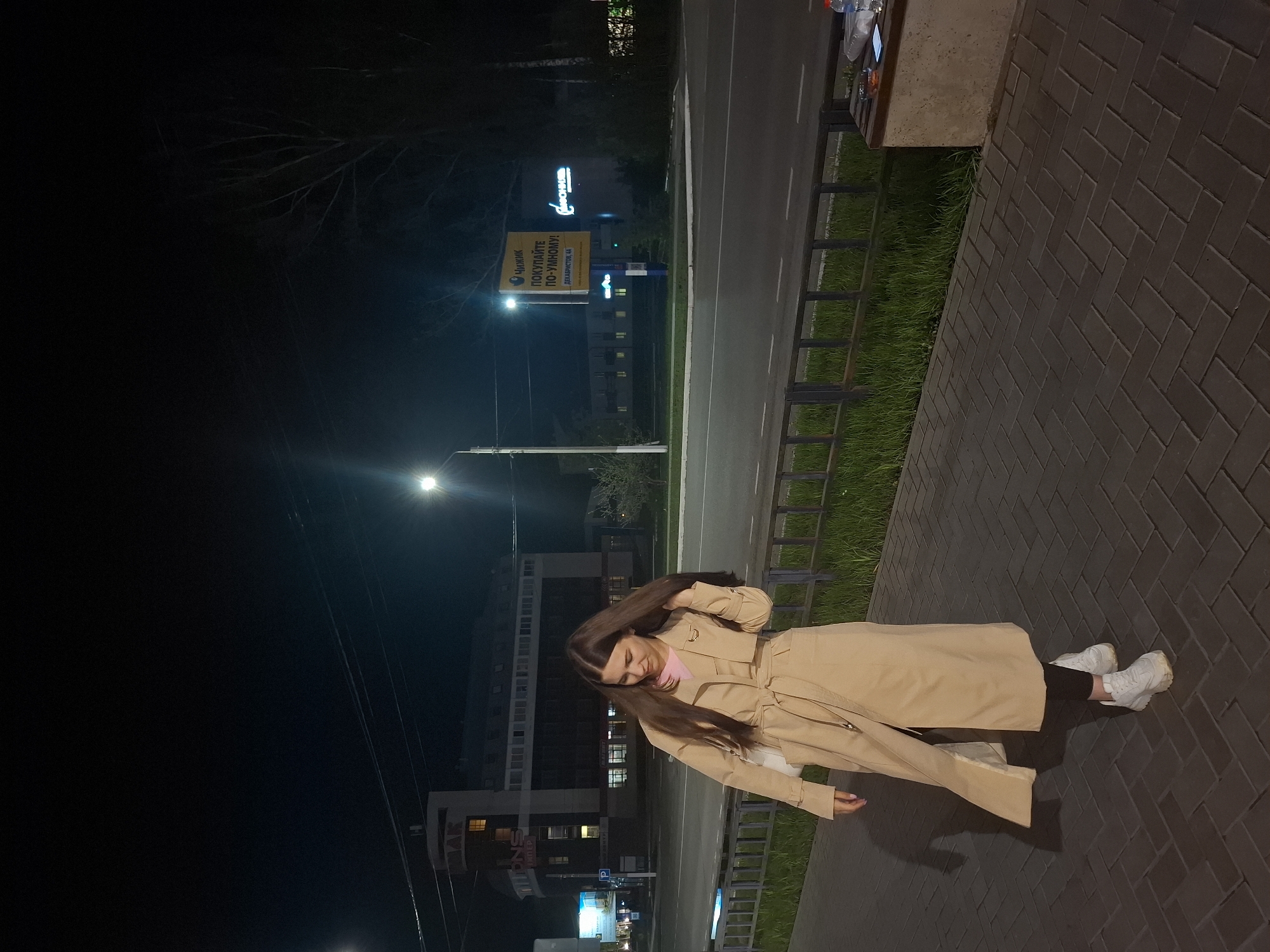 The image shows a woman standing on a brick-paved sidewalk at night in the city of Chaikovsky, Russia. She is wearing a light brown trench coat, black leggings, and white sneakers. Her long, dark hair is partially obscuring her face. Behind her is a street with a billboard visible, displaying text in Russian, "Чайник покупайте по-умному! Декабристов, 14" which translates to "Buy a kettle wisely! Dekabristov, 14". Further behind, a multi-story building with lighted windows is visible. Streetlights illuminate the area, and the sky is dark. Adjacent to the sidewalk is a narrow strip of grass and a low metal fence.