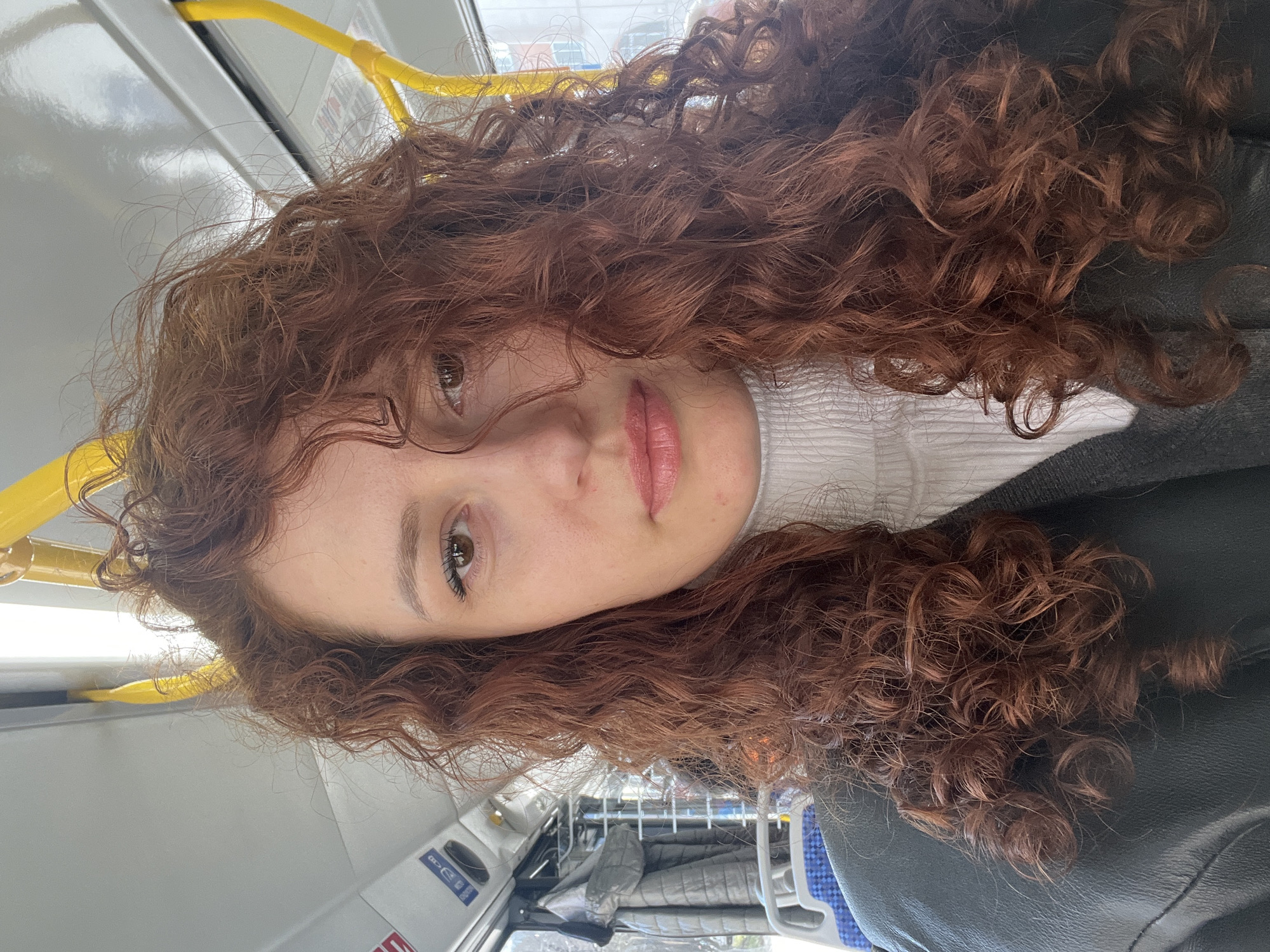 A young woman with auburn curly hair is in a bus or train. She has light skin, brown eyes, and light pink lipstick. She is wearing a white turtleneck sweater and a dark colored jacket. A yellow hand rail is visible above her head. The bus has various signage with pictograms and text on the side wall. A quilted jacket is on a seat across the aisle. There is nothing in the image to confirm the location is in Saint Petersburg, Russia.