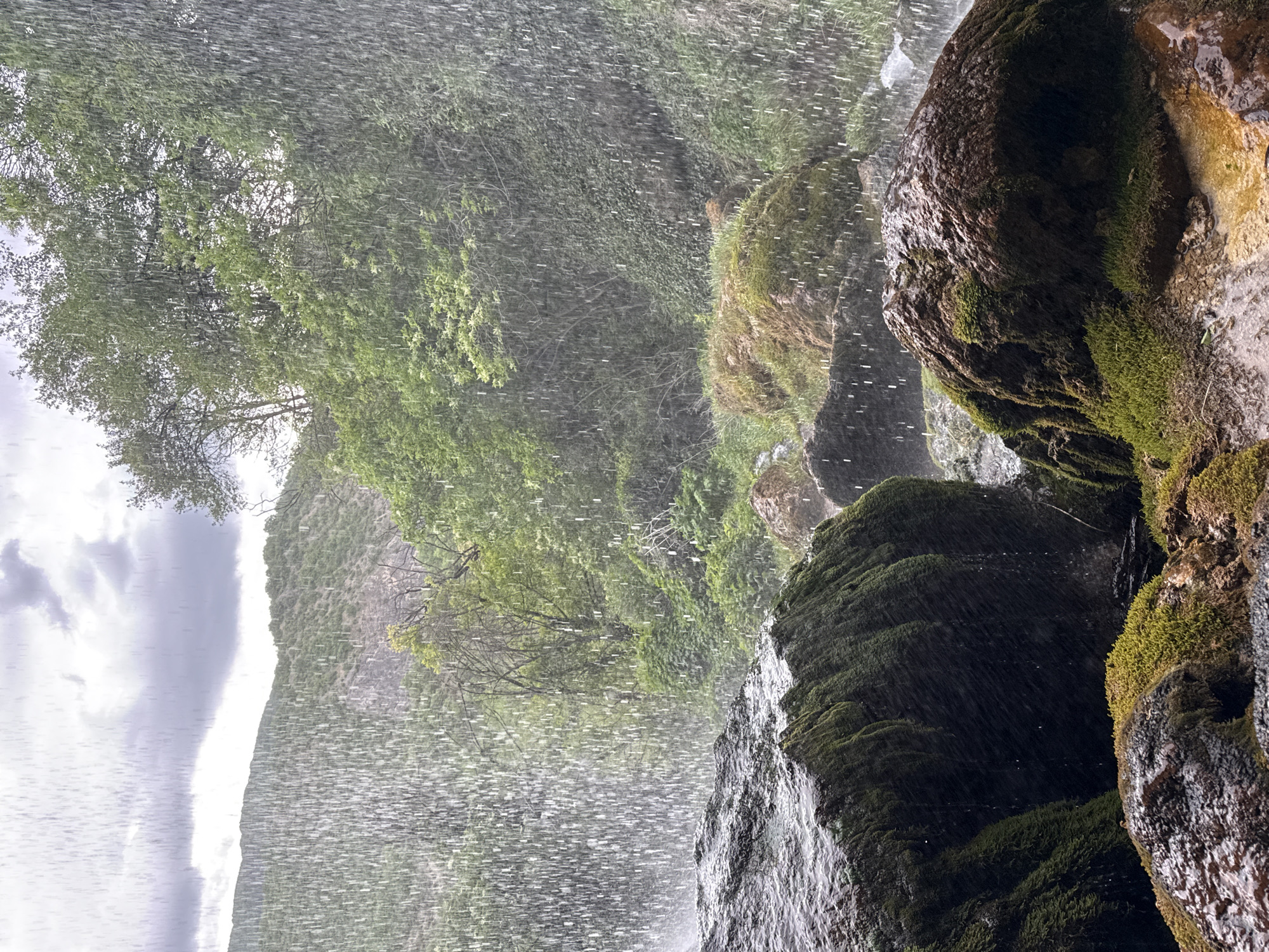 A landscape shot from behind a waterfall in Razhdavitsa, Bulgaria. The focus is on the curtain of water and the moss-covered rocks in the foreground. Behind the waterfall, a hillside covered in green trees is visible under a cloudy sky. The water creates a blurred effect, adding a sense of motion to the scene. The moss on the rocks appears wet and vibrant.