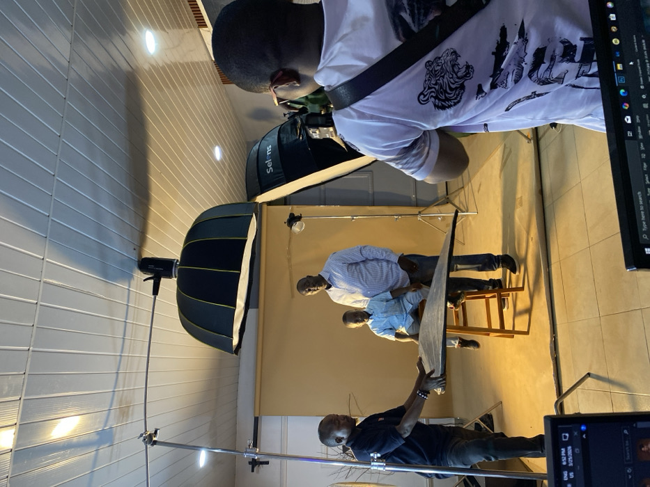 The image depicts an indoor photography studio in Bukuru, Nigeria, during an active photoshoot. In the center, a young boy wearing a light blue short-sleeved button-up shirt and dark pants is seated on a wooden stool, positioned next to an adult male in a light blue checkered long-sleeved shirt and dark pants; both subjects are posing for the camera. To the left, a male assistant in a dark blue polo shirt and dark pants holds a rectangular silver reflector, directing light towards the subjects. A male photographer, viewed from behind, wears a white t-shirt with dark tiger graphics and operates a DSLR camera, looking through its viewfinder.
The studio setup includes a tan seamless paper backdrop behind the subjects. Overhead and to the left, a large black softbox with a yellow interior stripe is mounted on a stand. Another large white softbox, bearing the "Selens" brand name, is positioned to the right of the subjects. A smaller light head on a stand is visible behind the adult male subject. The ceiling is white paneled with recessed lighting, and the floor consists of light-colored tiles. In the immediate foreground, two laptop screens are visible; one displays an interface with the time "6:52 PM" and date "3/25/202X".