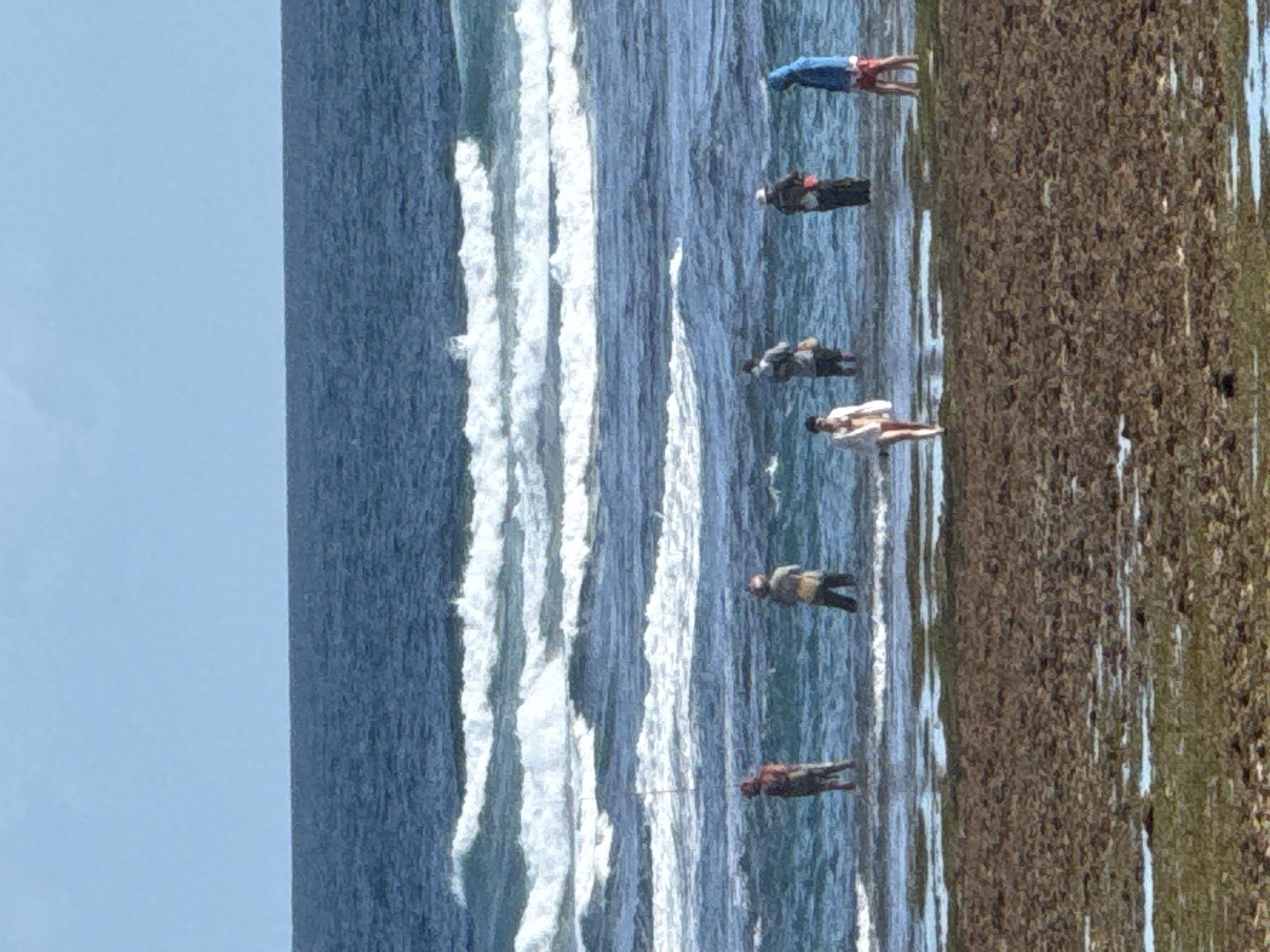 The photograph depicts a scene along the coastline of Kuta Selatan, Indonesia, featuring a beach, ocean, and several individuals engaged in fishing activities. Seven people are visible in the image, positioned along the shoreline. They are spaced out along the edge where the land meets the ocean, and appear to be in the act of fishing. The sea is showing evidence of wave action. The photograph is taken in daylight, with clear visibility.