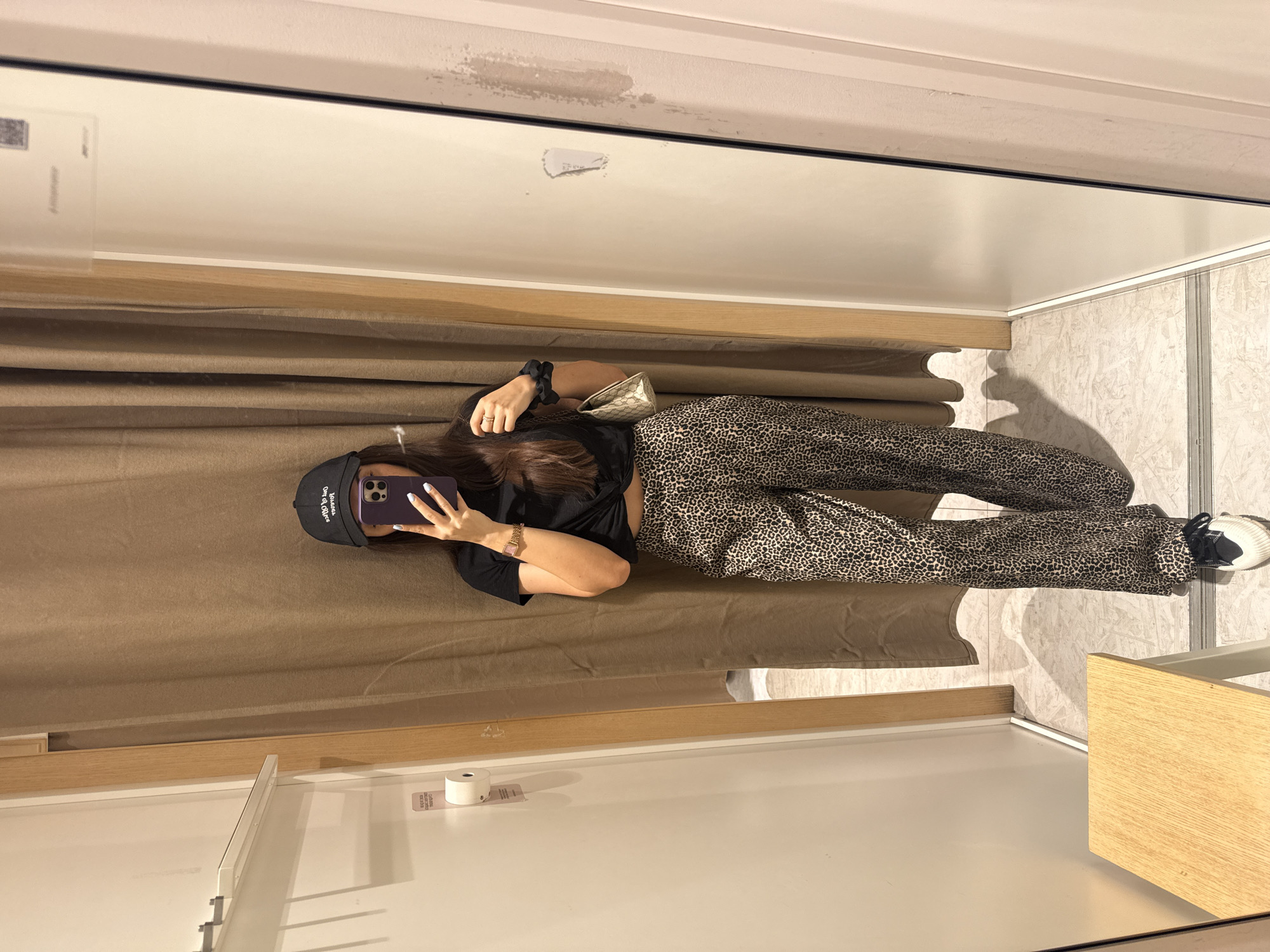 The image is a reflection of a person taking a selfie in what appears to be a fitting room. The person is wearing a black t-shirt, leopard print pants, a black cap, and is holding a smartphone. A small, patterned purse sits near her. The fitting room has a brown curtain and white walls with wooden trim. Based on the limited information, it's impossible to determine if the location is in Plovdiv, Bulgaria.