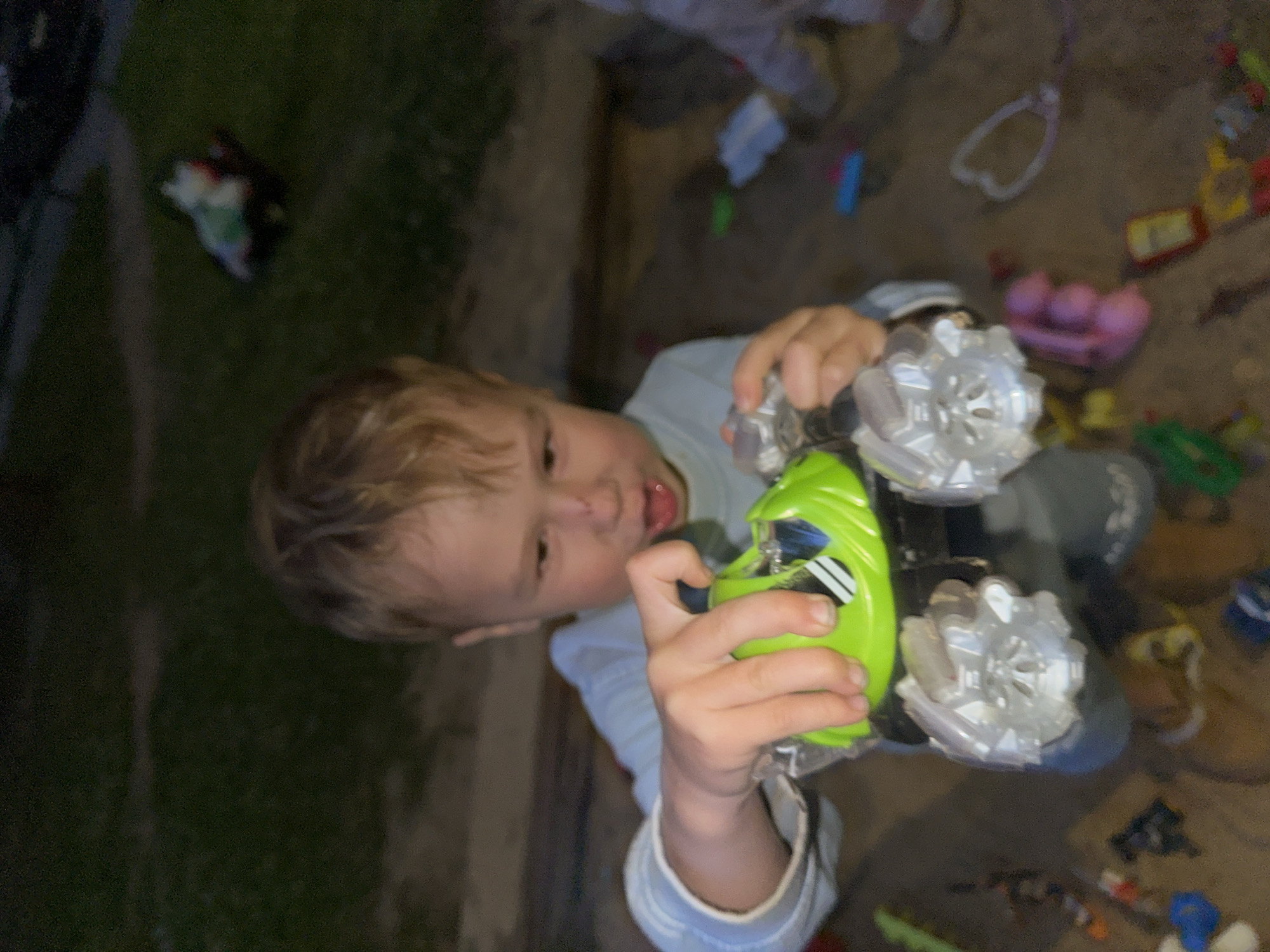This media file shows a young child holding a bright green toy car with large silver wheels. The child has blonde hair and is wearing a light blue long-sleeved shirt. The child's tongue is sticking out slightly, suggesting concentration or playfulness. The toy car has a black chassis and clear, intricate wheels that appear to be illuminated. The setting appears to be outdoors, possibly a backyard or sandbox, with sandy ground and patches of grass visible. Various other toys are scattered around, including a pink toy cradle with balls, a blue toy, and other colorful toy pieces. The lighting suggests it might be late afternoon or early evening. The overall mood is one of childhood play and exploration. There is no visible text in the image.