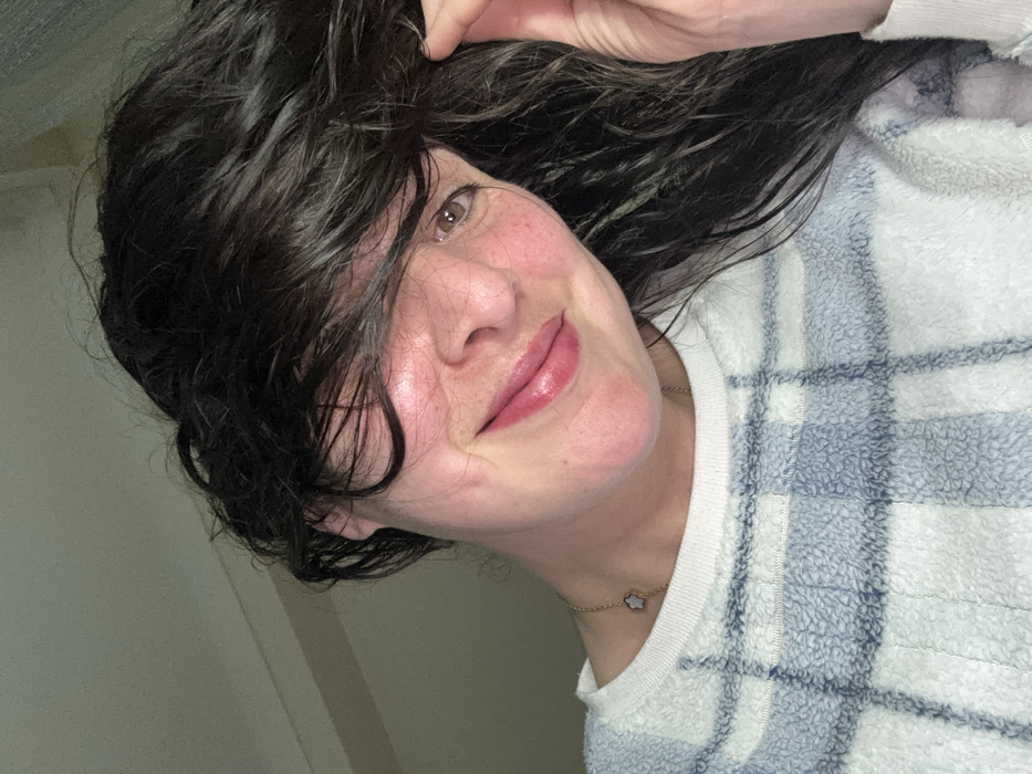 A person with dark, wet hair partially covering their face smiles. They are wearing a white and blue plaid sweater. Their skin has a slight redness, and they are wearing lip gloss. A gold necklace with a cloud-shaped pendant is visible. The background is a light, neutral color.