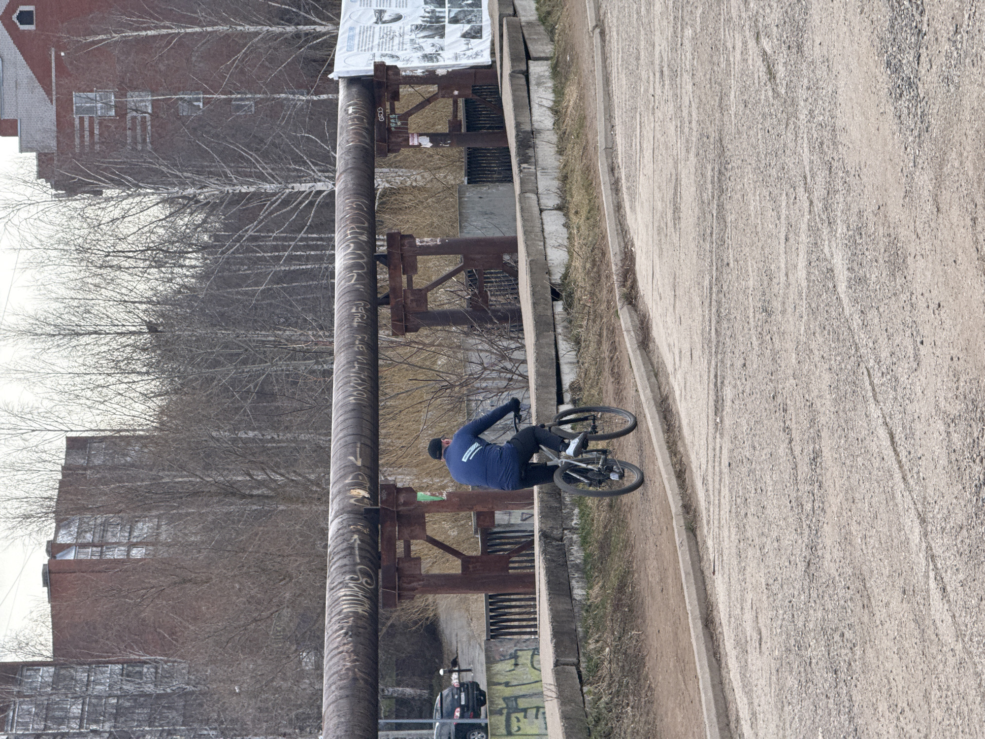 The image shows a man riding a bicycle on a concrete path. The location is Votkinsk, Russia, near a bridge. He is wearing a blue jacket, black pants, and a black cap. He's riding near a large rusty pipe with Cyrillic writing. The background includes bare trees, brick buildings, and a gray car. Graffiti is visible on the bridge's support. The path is made of light gray gravel and is bordered by a concrete curb.