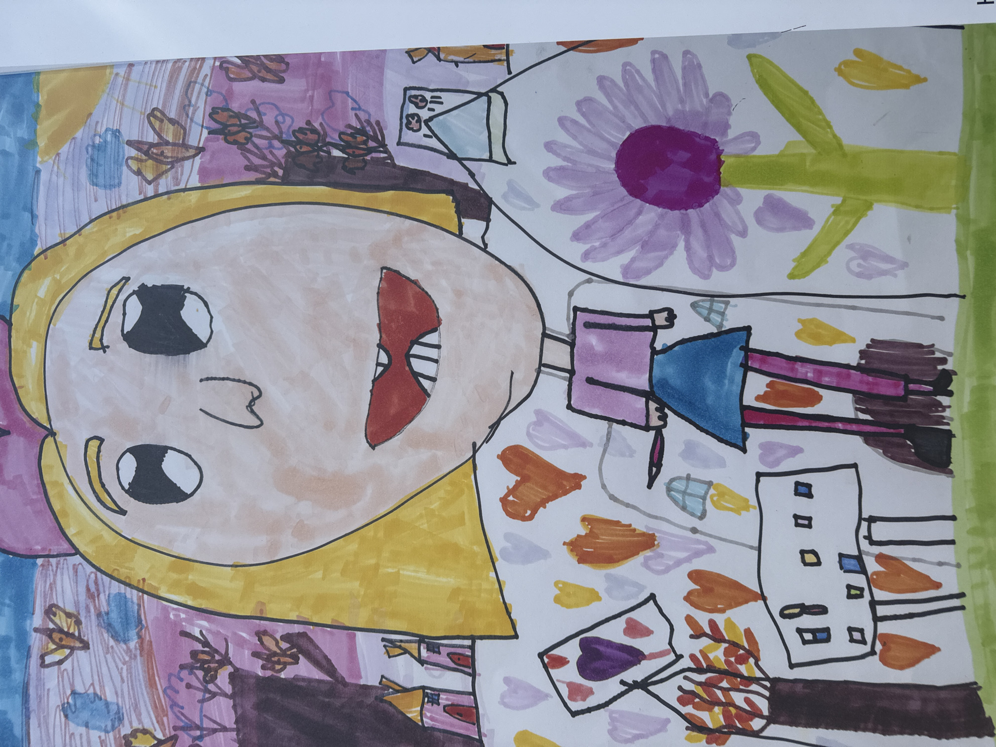 The image is a child's drawing featuring a large, cartoonish face with blonde hair, wide eyes, and a wide-open mouth displaying a red interior. The face is set against a colorful background filled with various shapes, patterns, and objects. These include butterflies, leaves in autumn colors, hearts, buildings, and a large purple flower.
The scene appears to be a cityscape as the buildings and city elements are present in the drawing. While precise location in Sofia is impossible to determine from the drawing, the subject of the drawing may have lived in Sofia, Bulgaria.