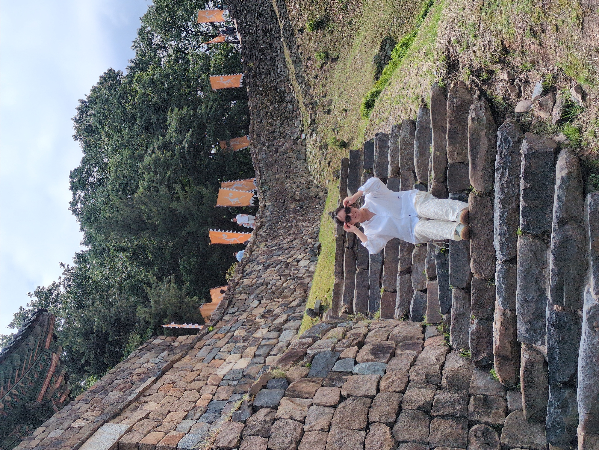 A medium, rotated shot shows a person on a set of stone stairs in front of a stone wall with orange flags hanging from it, likely in Gongju-si, South Korea.
The person, with dark hair pulled back and wearing sunglasses, a white collared shirt, and light pants, stands on the steps with both hands near their face, possibly adjusting their sunglasses.
The stairs are made of irregular, dark grey, stacked stones. Next to the stairs is a stone wall made of light brown and grey stones. Hanging from the wall are several orange flags with animal designs. A grassy slope rises behind the wall to the right.
Dense, green trees are to the left of the wall, under a slightly overcast sky. Part of a building's roof is visible near the bottom left corner.