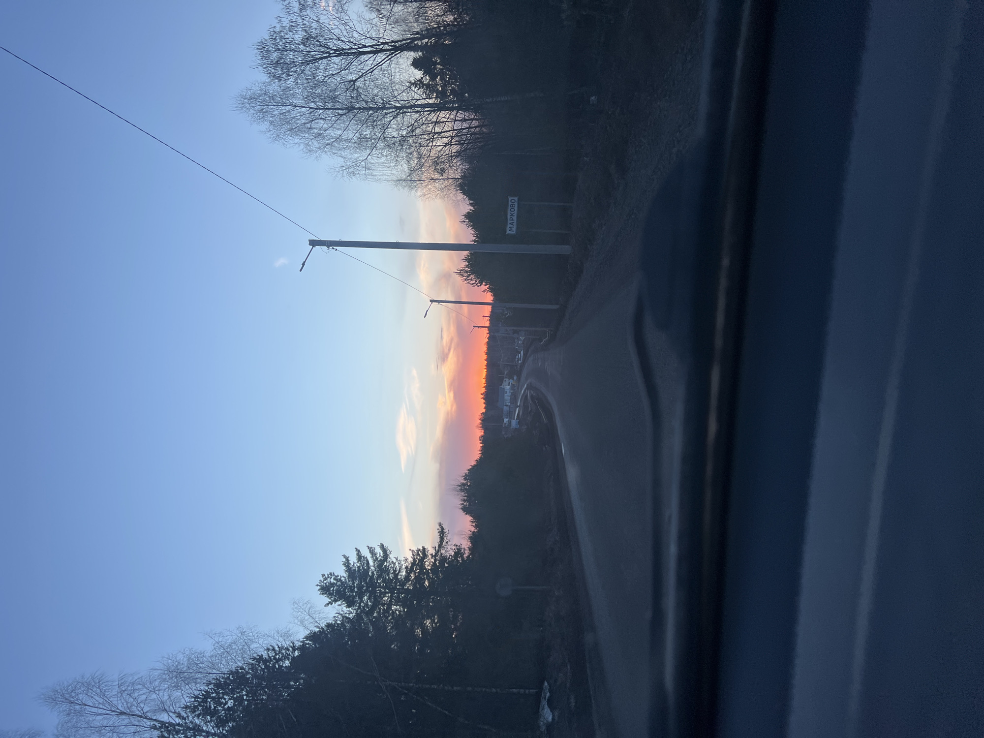 The image shows a road leading into Markovskii, Russia, indicated by a road sign. The view is likely from inside a vehicle. The sky displays colors of sunset or sunrise. Power lines run along the road. Trees and vegetation line both sides of the road. The light conditions suggest dusk or dawn.