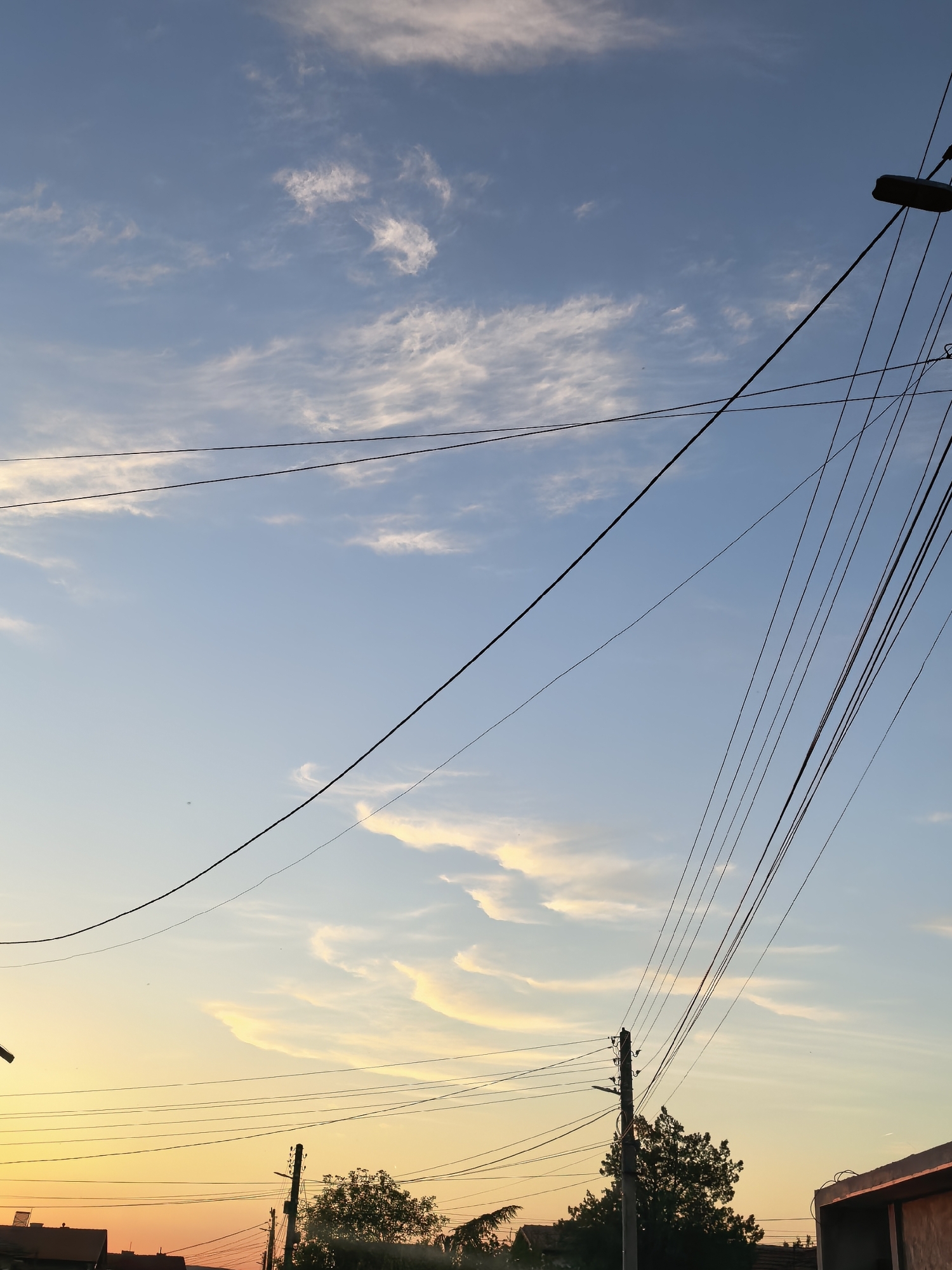 The image depicts a view towards the sky at what appears to be either sunrise or sunset in an urban environment, likely in Sapareva Banya, Bulgaria, given the prompt's instruction. The sky transitions from a pale blue at the top of the frame to orange and yellow hues near the horizon. Several layers of clouds are visible, with elongated formations near the horizon and scattered, wispy clouds higher up. Power lines are strung across the scene, obscuring parts of the sky. Utility poles are also visible, with trees, rooftops, and the corner of a building suggesting a residential area. A streetlight is present on the upper right. No people are seen.