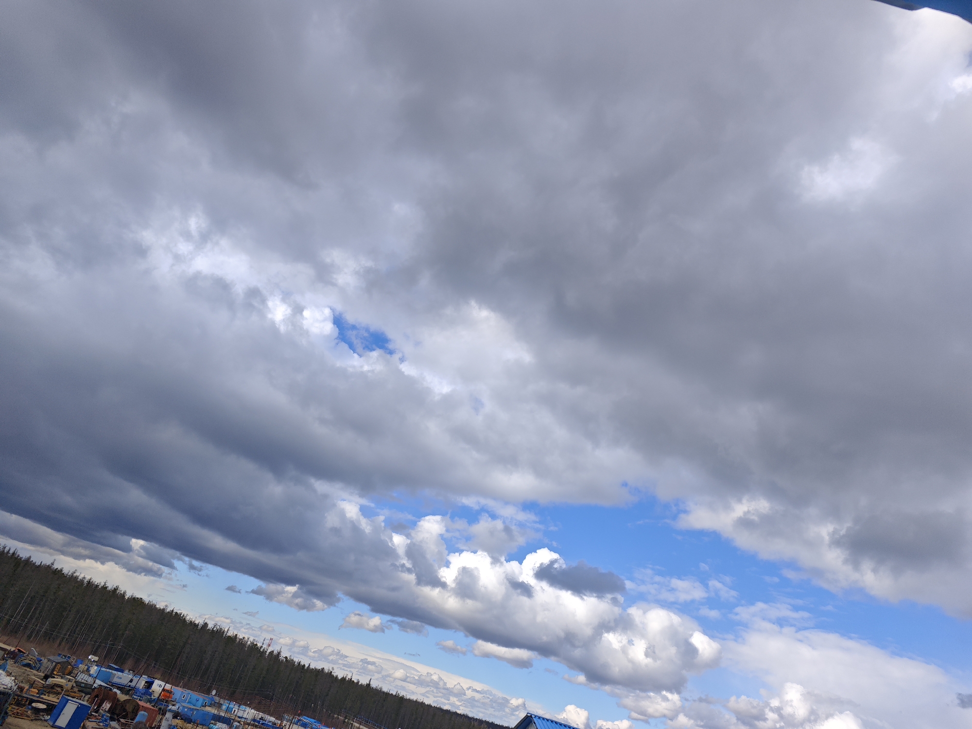 The image depicts a cloudy sky over a location in Unknown, Russia. The sky is dominated by gray and white clouds, with patches of blue sky visible in between. Below the sky, there is a line of trees and some buildings or structures. The overall scene appears to be outdoors.